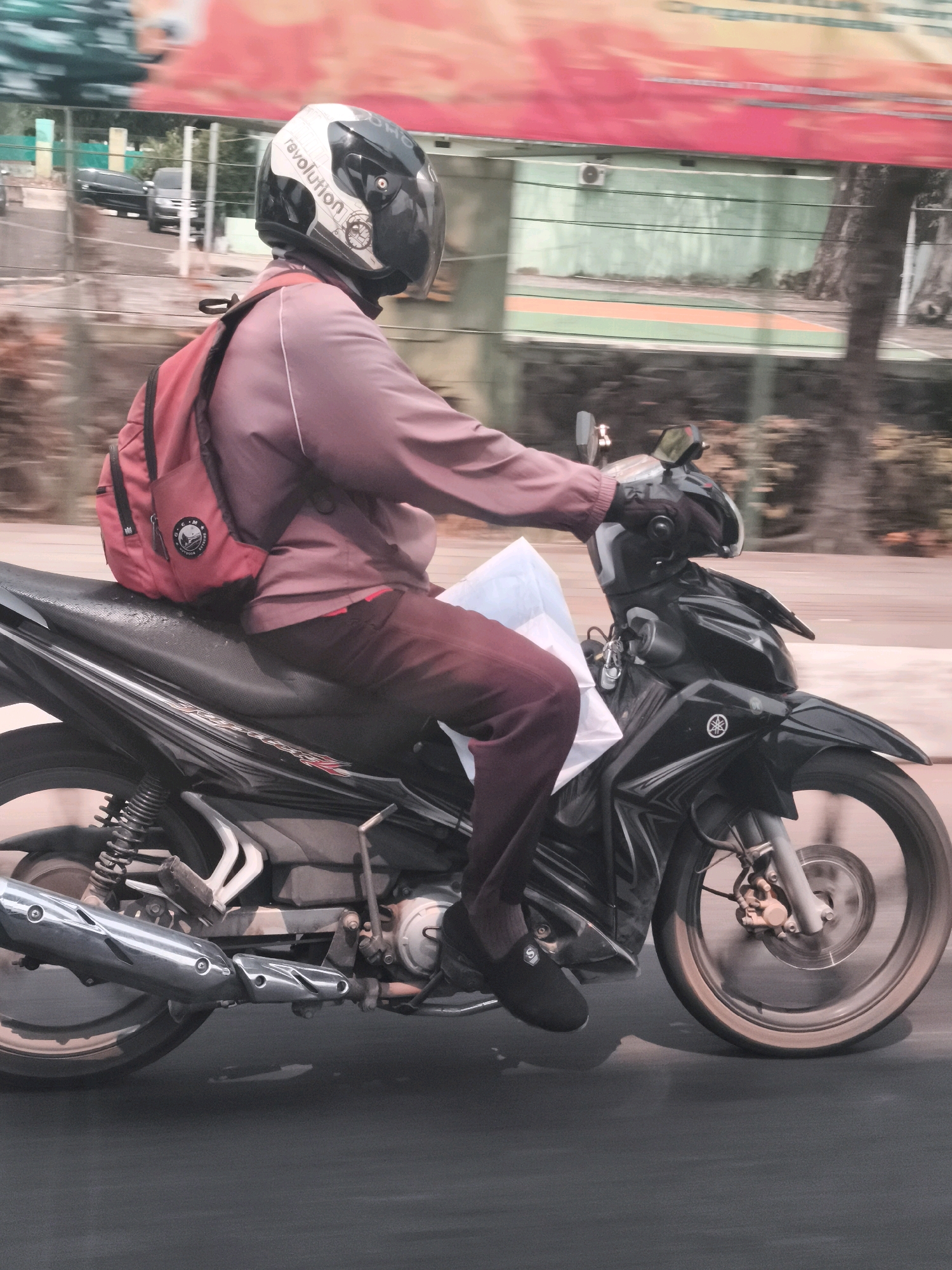 A person wearing a red backpack and a helmet is riding a black motorcycle. The rider is wearing a long-sleeved shirt and pants. The motorcycle has a black body and silver accents. The scene appears to be in an urban environment, possibly in Palembang, Indonesia, as indicated by the background which includes a building with green walls and a sports court. A large red and orange banner is visible in the upper background, suggesting a commercial or public area. The image is taken from a side view, capturing the motion of the motorcycle.