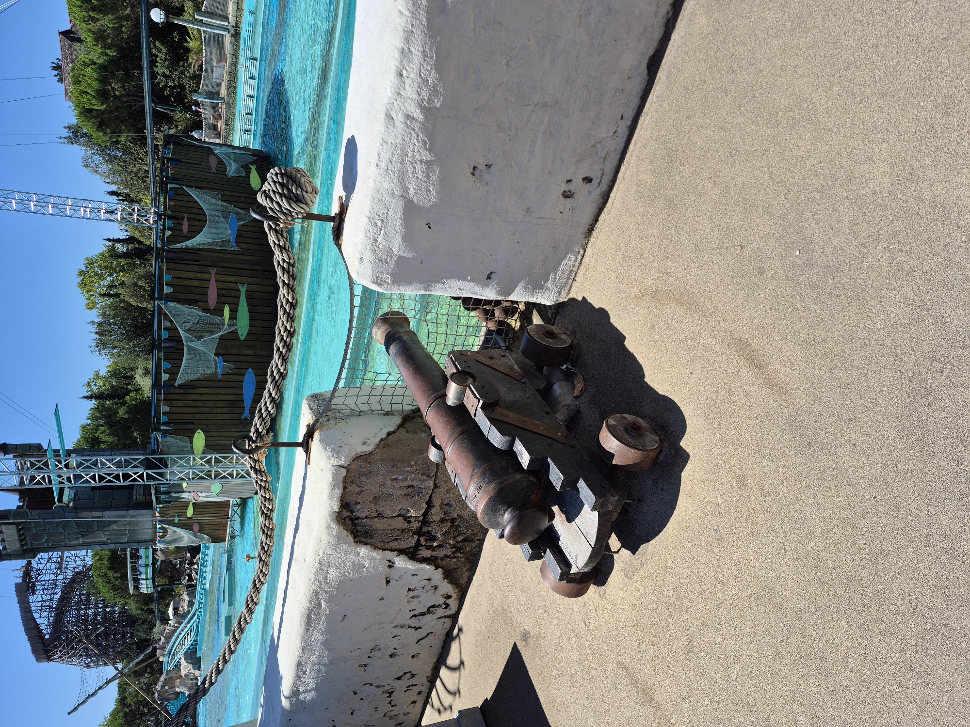 In Rust, Germany, on a sunny day, a replica of an antique cannon sits on a sandy path next to a white, weathered concrete barrier. The cannon, made of metal and wood, is positioned in front of a large body of turquoise water, which appears to be part of an amusement park attraction. In the background, a wooden ship facade with netting and colorful fish decorations is visible, along with a tall structure resembling a roller coaster and a metal tower. The scene suggests a themed area within an entertainment venue, likely a water park or amusement park. No people are prominently featured or interacting with the cannon or the environment. The overall atmosphere is recreational and vibrant, characterized by bright sunlight and clear blue skies.