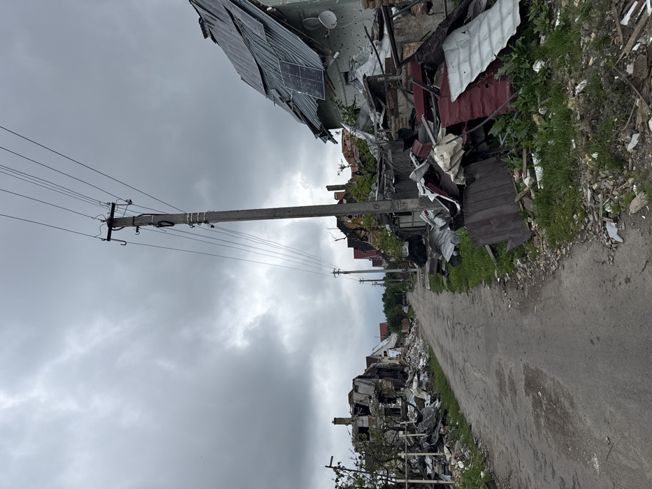 A street with buildings destroyed. On the left, the remnants of a building with solar panels hang precariously. Debris litters the landscape, with roofs and structural elements scattered. A gray sky provides a somber backdrop. Power lines run along concrete poles. Vegetation is present along the roadside. The road is paved and leads into the distance.