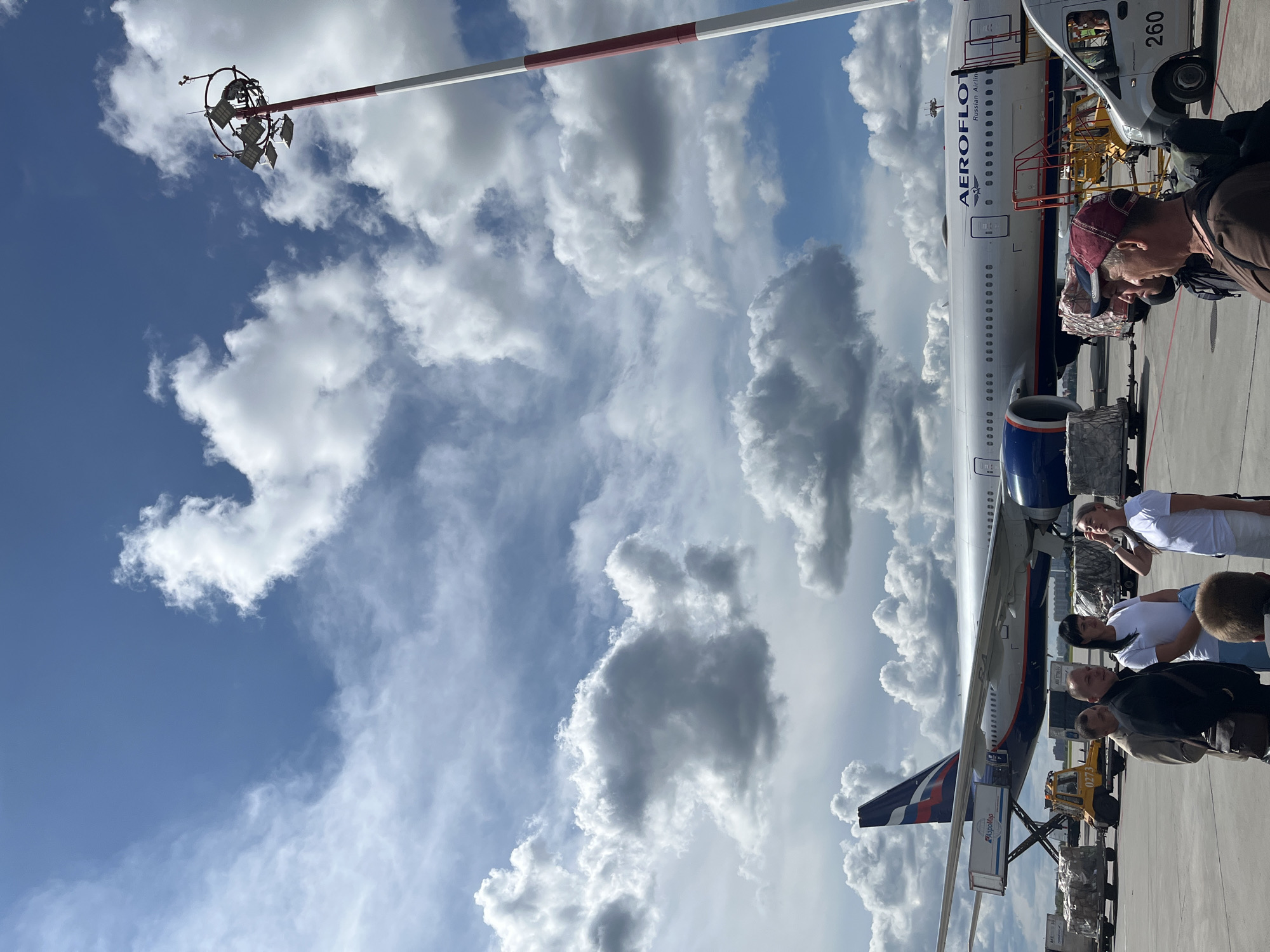 This is an eye-level, medium shot, taken outdoors on an airport tarmac under a partly cloudy sky. In the background, a large Aeroflot airplane is parked, with its tail fin featuring the Russian flag and Aeroflot logo visible on the left. The plane has "AEROFLOT Russian Airlines" written on its fuselage. To the right of the plane is a tall light pole with multiple lights on top, painted with red and white stripes.
In the foreground, a group of six people are standing on the tarmac. On the far left, three men are standing together, one wearing a beige jacket and a dark bag slung over his shoulder. Next to him, a man in a black jacket and dark pants is facing the right. To his right, a woman in a white t-shirt and light blue pants is looking towards the man in the black jacket. Beside her, a woman in a white t-shirt is looking to her right, with her hand raised to her forehead. In the front right of the frame, a man wearing a red baseball cap is facing the left, and another person's head and shoulders are visible in the extreme foreground.
Various airport equipment is scattered around the tarmac, including cargo containers, a yellow tow tractor, and a white van with the number "260" visible on its rear door. The scene appears to be taking place during the daytime, with bright sunlight casting shadows. The airport is located in Khimki, Russia, a suburb of Moscow, known for hosting Sheremetyevo International Airport.