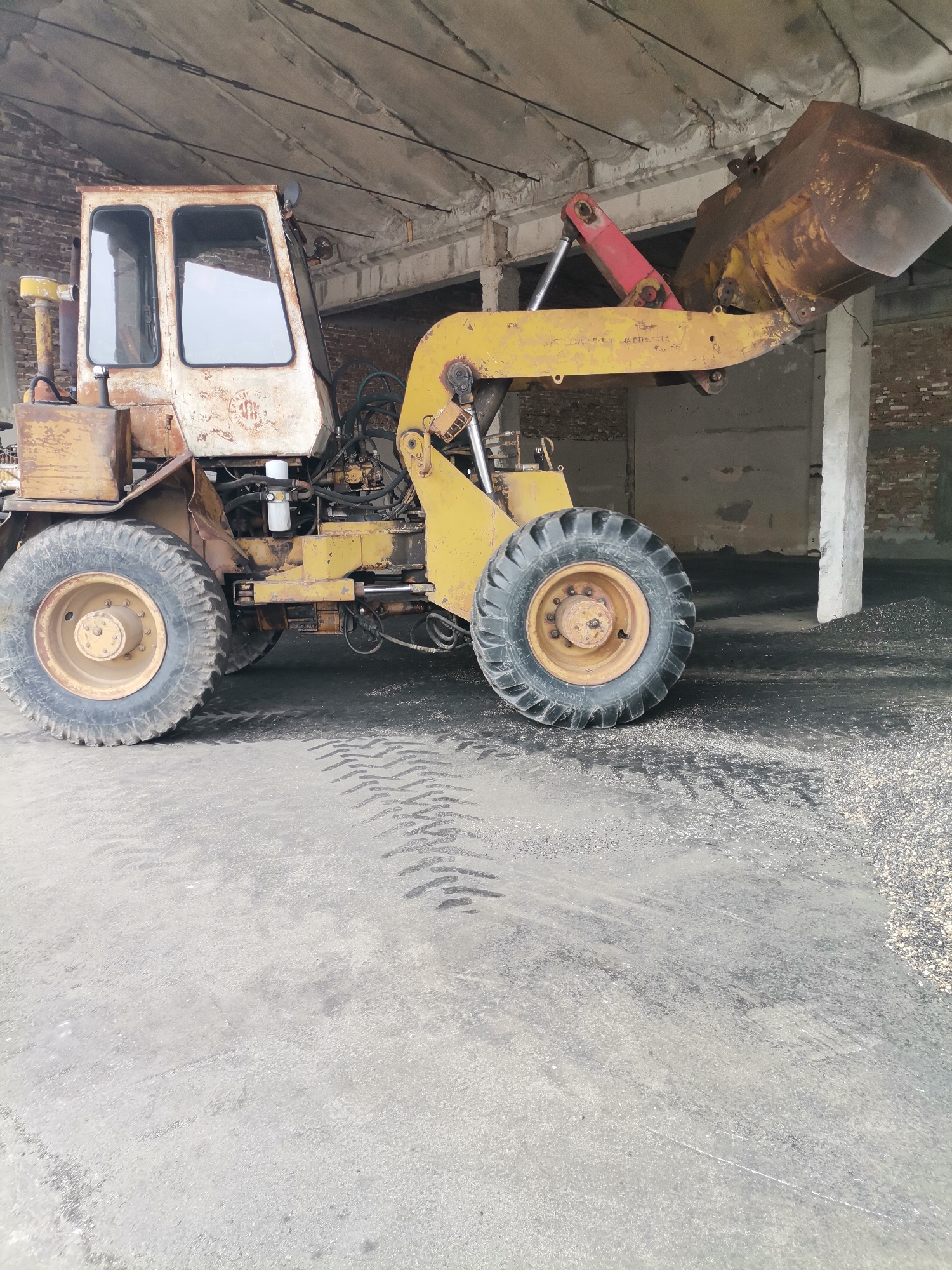 The image displays a weathered yellow front-end loader, also known as a wheel loader, parked inside what appears to be an aged industrial or agricultural building in Kochovo, Bulgaria. The machine exhibits significant wear, with rust and faded paint across its body. Its large, rusty bucket is elevated and filled with a dark, granular material, possibly coal, aggregate, or a similar bulk commodity.
The loader's cab is an off-white color, also rusty, featuring two prominent windows. Beneath the cab, various mechanical components, including hydraulic hoses, cylinders, and a visible white oil filter, are exposed. A section of the hydraulic arm connecting to the bucket is painted red. The machine's large, heavy-duty tires have aggressive tread patterns, and clear tire marks are visible on the concrete floor, suggesting recent activity. The right rear tire is partially surrounded by a pile of the same dark granular material.
The setting is a large, somewhat dilapidated indoor space with a concrete floor. The walls are a mix of exposed brickwork and concrete. The concrete ceiling shows visible structural elements, including beams and what appear to be rebar or tensioning cables. A white concrete support column stands on the right side of the frame. The overall scene suggests an active but old storage or processing facility.
No people are visible. The loader is stationary, implying it has either just completed or is about to undertake material handling. The time of day appears to be daytime, indicated by the ambient light filtering into the building. Visible text on the loader's cab includes a faint, circular, and illegible logo. Some unreadable markings are also present on the yellow boom arm.