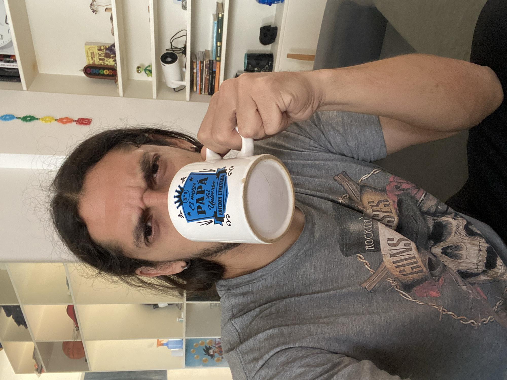 A man with long dark hair and an earring in his left ear is holding a white mug with a blue and white design, possibly related to Father's Day, in front of his face. He is wearing a gray Guns N' Roses t-shirt. The background shows a white bookshelf with various items, including books, a clock, a security camera, and decorative items. The man appears to be indoors, possibly in a living room or home office. The exact location within the city of Buenos Aires, Argentina, is indeterminable based on the provided image.