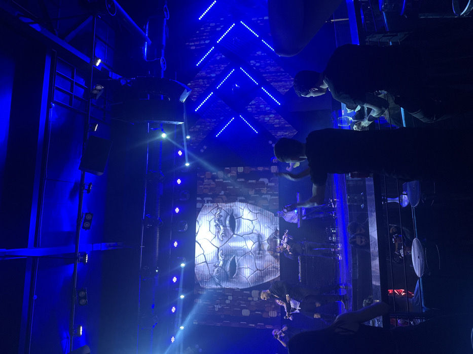 This image portrays the interior of a nightclub or music venue located in Varna, Bulgaria, illuminated predominantly by blue stage lighting. A stage occupies the background, featuring a large LED screen displaying a fractured, white mask resembling classical statuary. On the stage, a musical ensemble is performing, consisting of a central male performer in dark attire holding a microphone, a female individual in a light-colored top, a male playing a trumpet-like instrument, two male violinists, a male seated at a keyboard, and another male playing a stringed instrument resembling a guitar. To the right of the LED screen, a wall panel with diagonal blue LED light strips contributes to the visual effects. In the mid-ground, two male patrons are visible from behind. The male on the left, wearing a dark t-shirt, is captured with both hands raised in a clapping gesture. The male on the right, also in a dark t-shirt, holds a clear glass containing a beverage. Several shisha pipes are present on tables in the foreground, and additional indistinct audience members are visible in the dimly lit areas surrounding the seating. Overhead, structural elements, stage lights, and large speakers are visible against the dark ceiling.