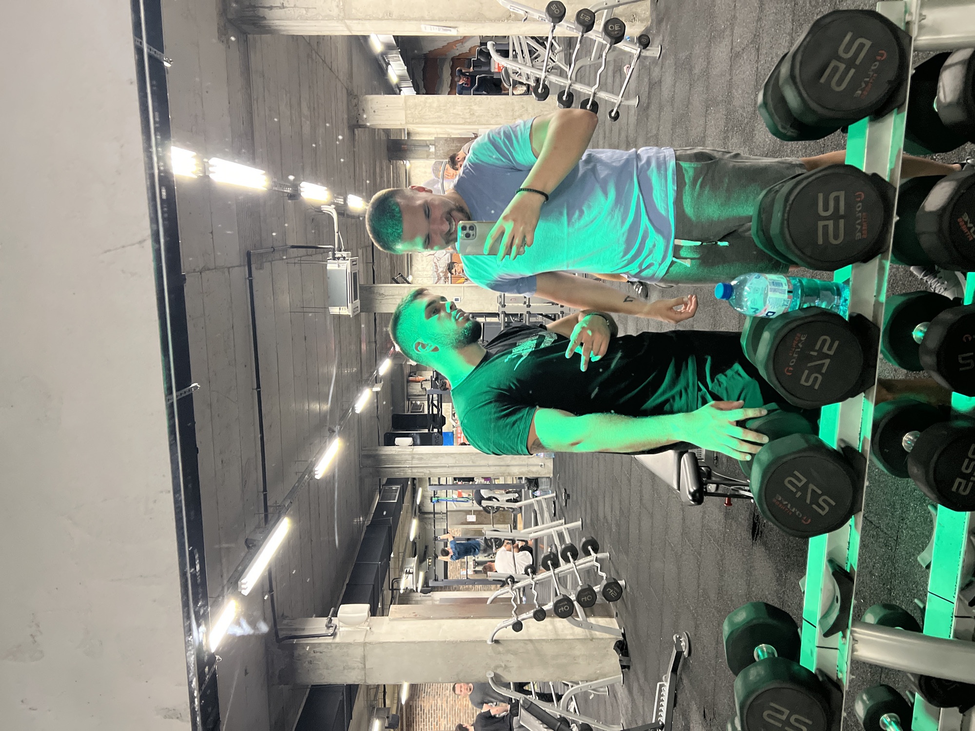 This photo, likely taken in a gym in Sofia, Bulgaria, shows two men in front of a mirror. The man closer to the mirror is holding a smartphone, seemingly taking a selfie with the other man. The man taking the photo is wearing a light blue t-shirt and gray sweatpants, while the other man is wearing a black t-shirt. Between them is a rack of dumbbells ranging from 2.5 to 52. Next to the dumbbells is a clear bottle of water. The gym itself has a utilitarian feel, with concrete columns and exposed lighting fixtures. Other gym equipment is visible in the background, including weight machines and additional dumbbell racks. Other people can be seen in the background using the gym equipment. The overall lighting in the gym is bright.
