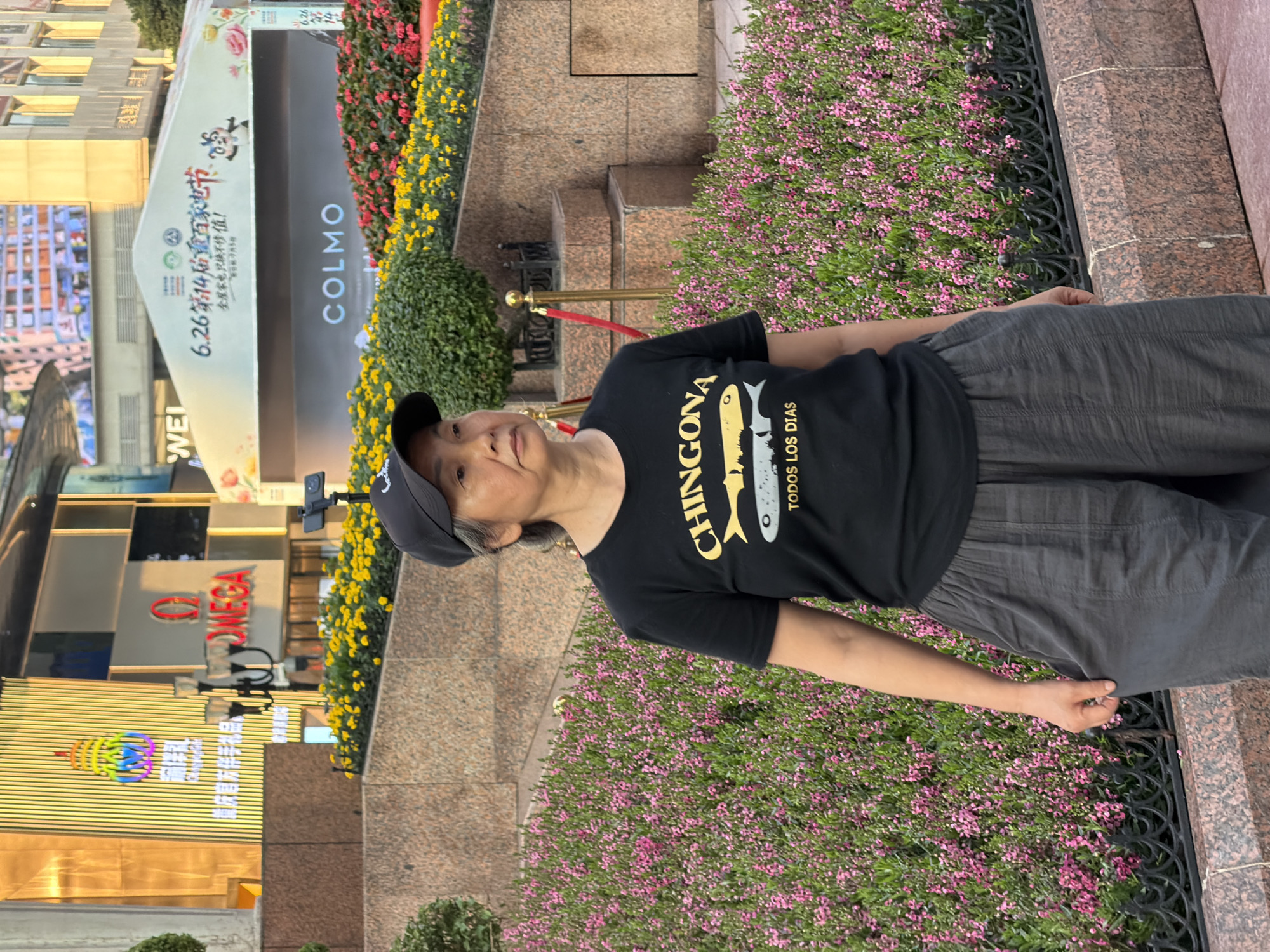 A middle-aged Asian woman wearing a black t-shirt and gray pants stands outdoors, facing right. The t-shirt has yellow text and a graphic of two fish. She is wearing a black baseball cap. Behind her, a multi-tiered landscape features yellow and pink flowers, with buildings and signage in the background. One sign displays "OMEGA" in red letters. Another sign in the distance advertises "6.26第14届重庆购物节" and "COLMO". The woman is positioned in front of a low, decorative black metal fence. The scene appears to be a public urban area in Yuzhong District, China.