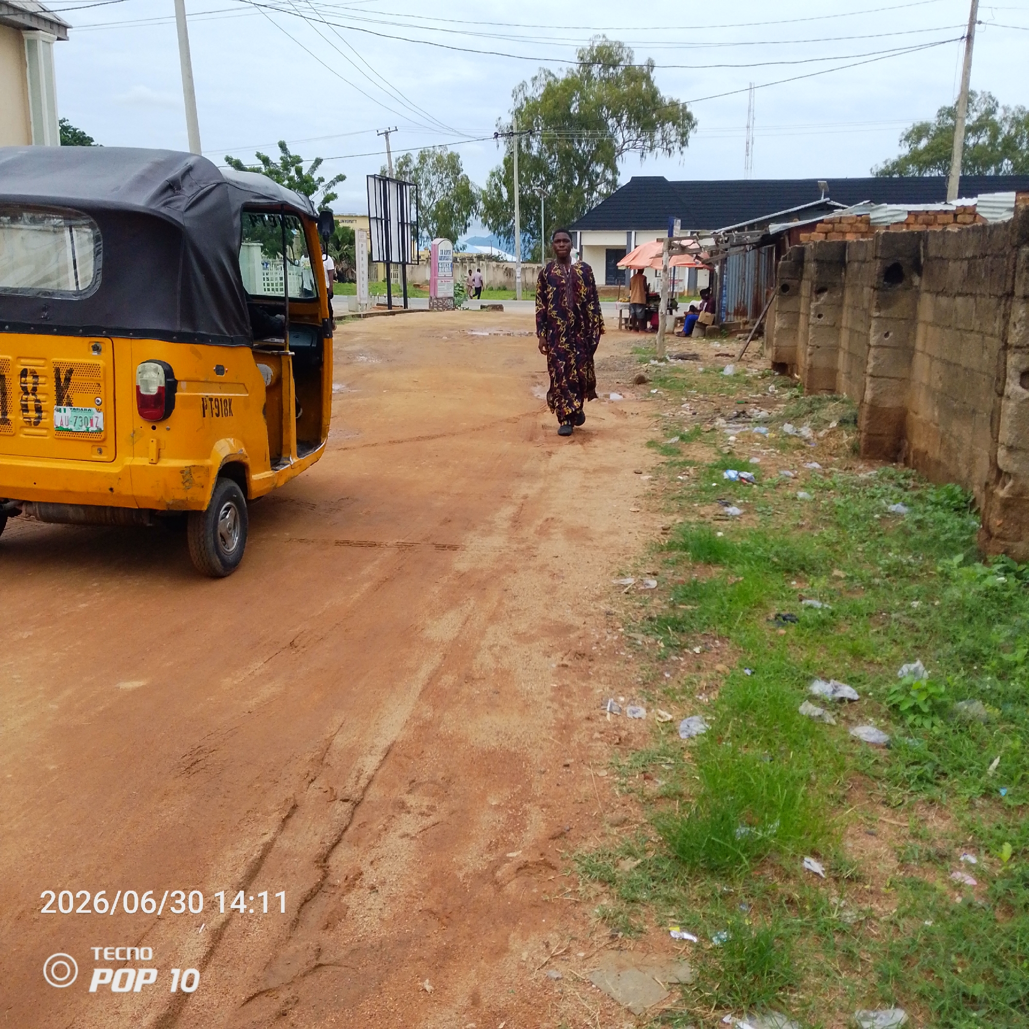 The image depicts an outdoor scene in Jalingo, Nigeria. In the foreground, a yellow, three-wheeled auto-rickshaw, commonly known as a "Keke Napep," is parked on a dirt road. The auto-rickshaw has a black canopy and a license plate that reads "LAU-730WZ." A person wearing a traditional, patterned robe is walking away from the viewer on the dirt road.
In the background, there are various structures, including buildings with dark roofs and a stone wall. Small stalls with orange canopies are visible, suggesting a market or roadside vendors. Several trees are scattered throughout the scene, and utility poles with overhead wires are prominent. The ground is a mix of reddish-brown dirt and patches of green grass, with some scattered litter, including plastic bags. The sky is overcast, indicating a cloudy or possibly rainy day.
The overall impression is one of a typical day in a Nigerian town, with everyday activities and common modes of transportation. The presence of the auto-rickshaw, the traditional clothing, and the architecture provide strong location cues for Jalingo, Nigeria. The date and time stamp at the bottom left corner of the image reads "2026/06/30 14:11" and the text "TECNO POP 10" is also visible.