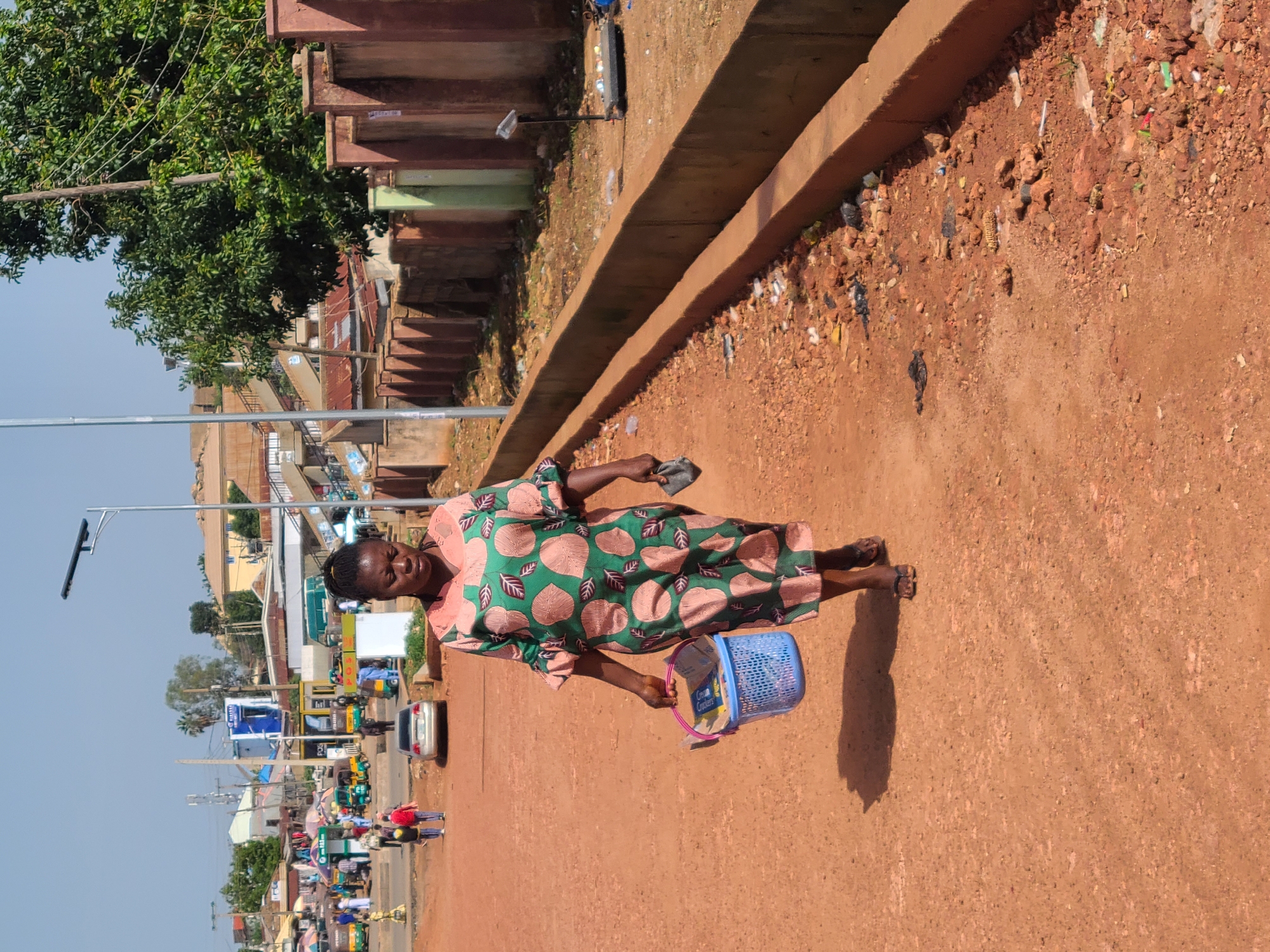 A Black woman walks on a dirt road in Gura Top, Nigeria. She is wearing a green and pink patterned dress and carrying a blue plastic basket in her left hand and a dark cloth in her right hand. The road is reddish-brown and dusty, with some loose stones and debris scattered along the edges. In the background, there are buildings, market stalls, and various vehicles, including a silver car, tricycles, and a green bus. A solar-powered street light is visible on the left side of the image, and trees and a concrete wall are present on the right. The sky is clear and blue, indicating a sunny day.