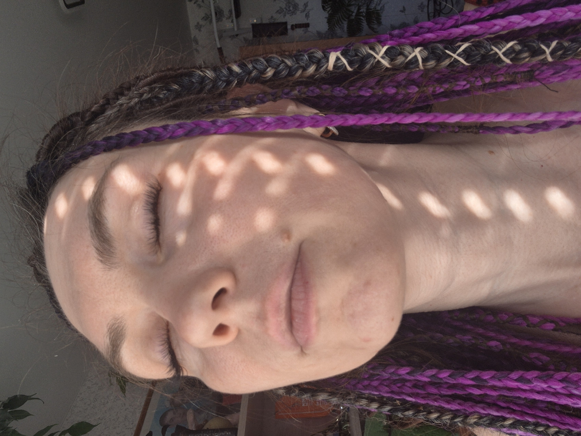 The image shows a close-up of a fair-skinned person's face in a vertical orientation. Their eyes are closed, and light filters across their face, creating a pattern of dappled shadows. The person has braided hair, with purple and mixed-color braids visible around their face. The background appears to be an indoor setting, with blurred objects and a plant visible in the lower part of the image, and a curtain with a floral pattern is visible at the top. The image is not easily identifiable as being in a specific city or location. The lack of clear landmarks or contextual clues makes it difficult to determine any further information about the scene.