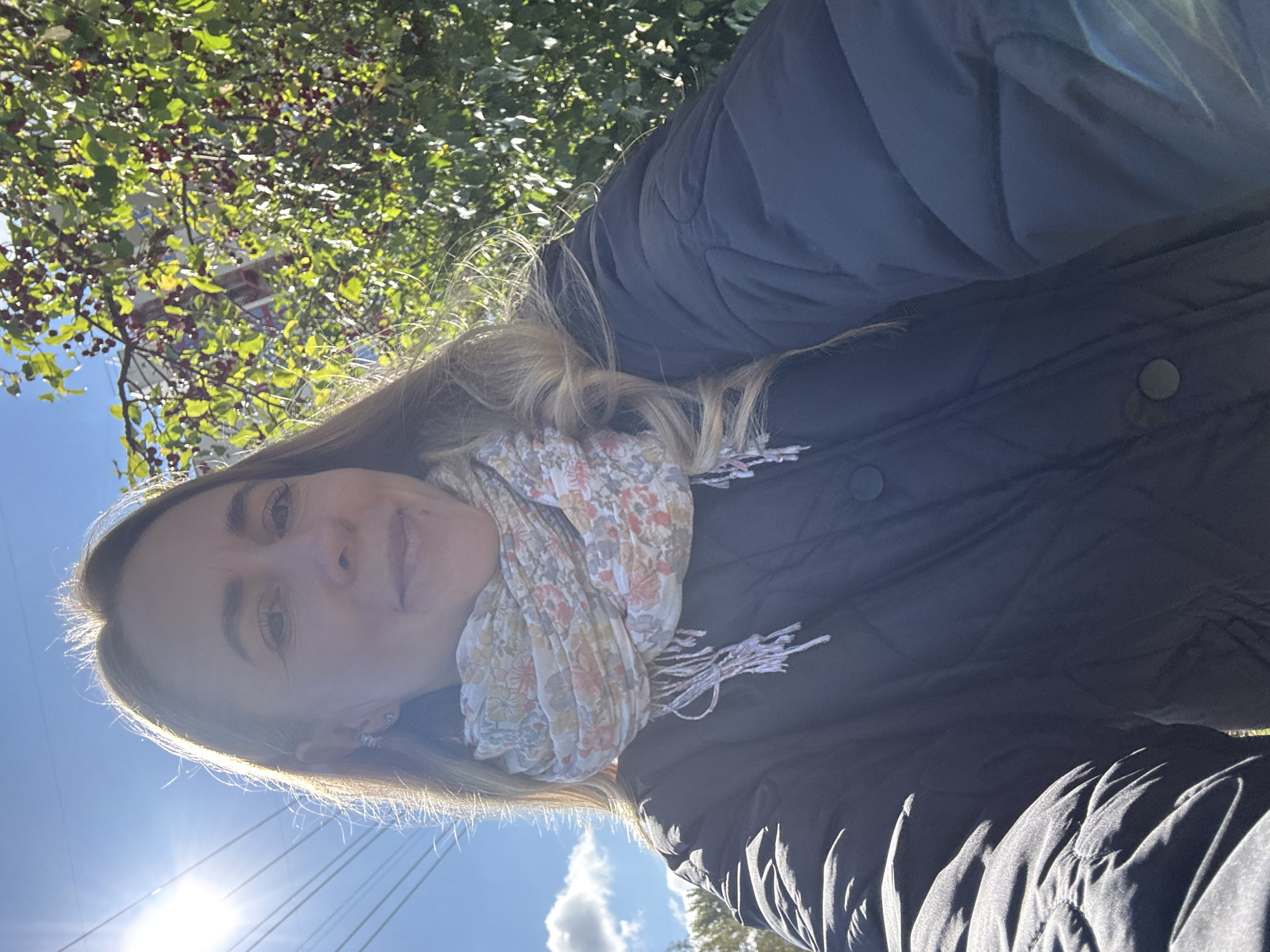The image is a selfie taken outdoors on a sunny day. A woman with blonde hair is in the foreground, wearing a black quilted jacket and a floral scarf. She is looking towards the camera with a slight smile. Behind her, a tree with red berries and green leaves is visible. In the background, there are power lines and a bright blue sky with some clouds. The scene appears to be in a residential area, likely in the city of Chaikovsky, Russia, given the presence of typical urban elements like power lines and trees.