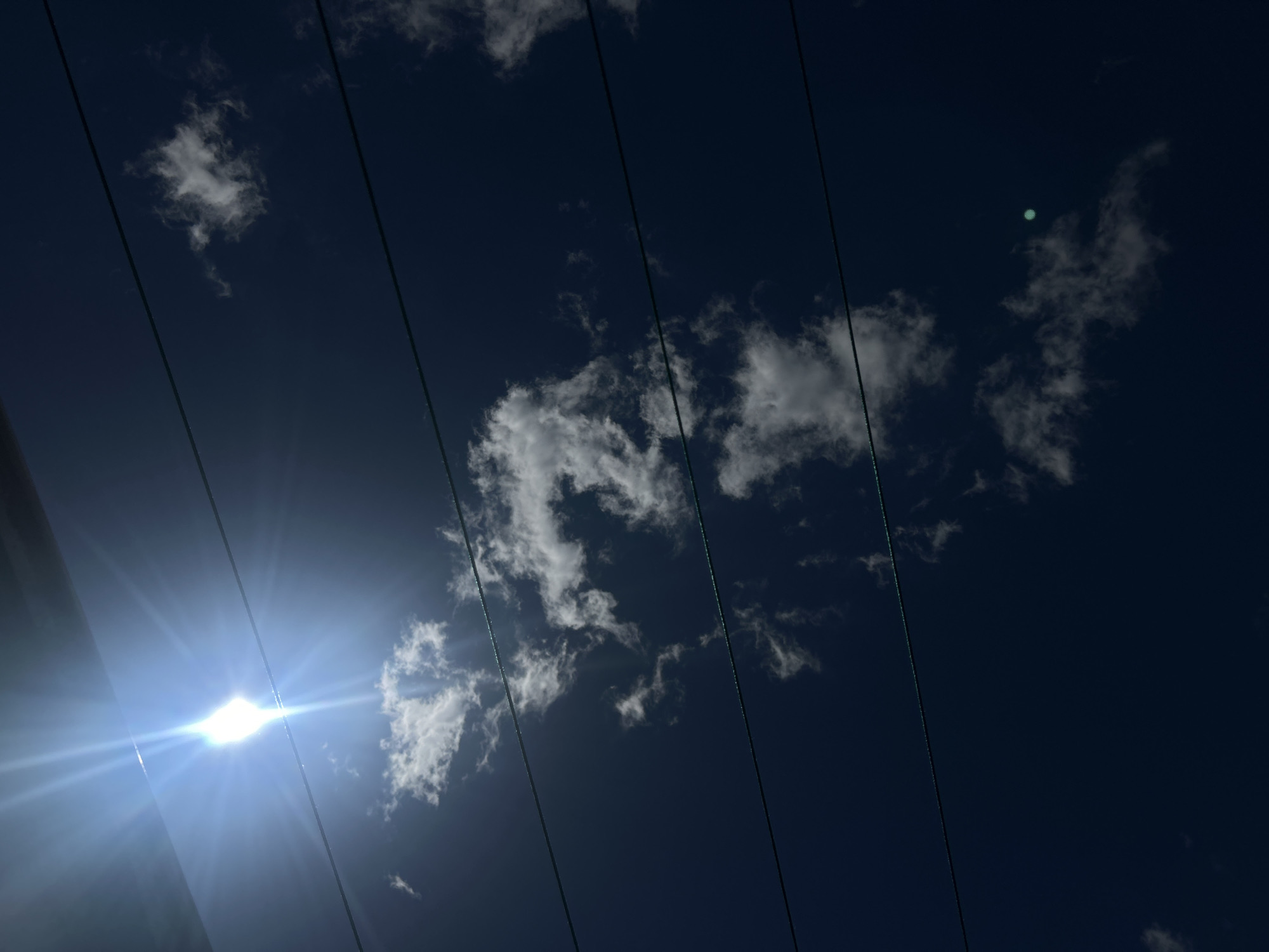 The image displays a sky scene with visible power lines crossing horizontally. The sun is brightly shining, creating a lens flare effect in the upper left portion of the frame. Wispy clouds are scattered across the dark blue sky. The lower edge of an unseen structure, possibly a building or overhang, is visible at the very top of the image. The perspective is looking upwards. No people or specific landmarks of Shumen, Bulgaria are identifiable in the provided image.