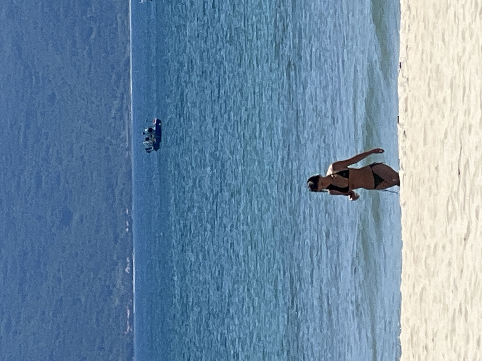 A person wearing a black bikini walks out of the ocean onto a sandy beach. The person is facing slightly away from the camera. The water is blue and calm, with small waves. In the distance, a small boat with several people on board is visible. Behind the water, a lush green mountain range dominates the background. The scene appears to be a beach in Da Nang, Vietnam, given the characteristic scenery of coastal mountains meeting the sea.