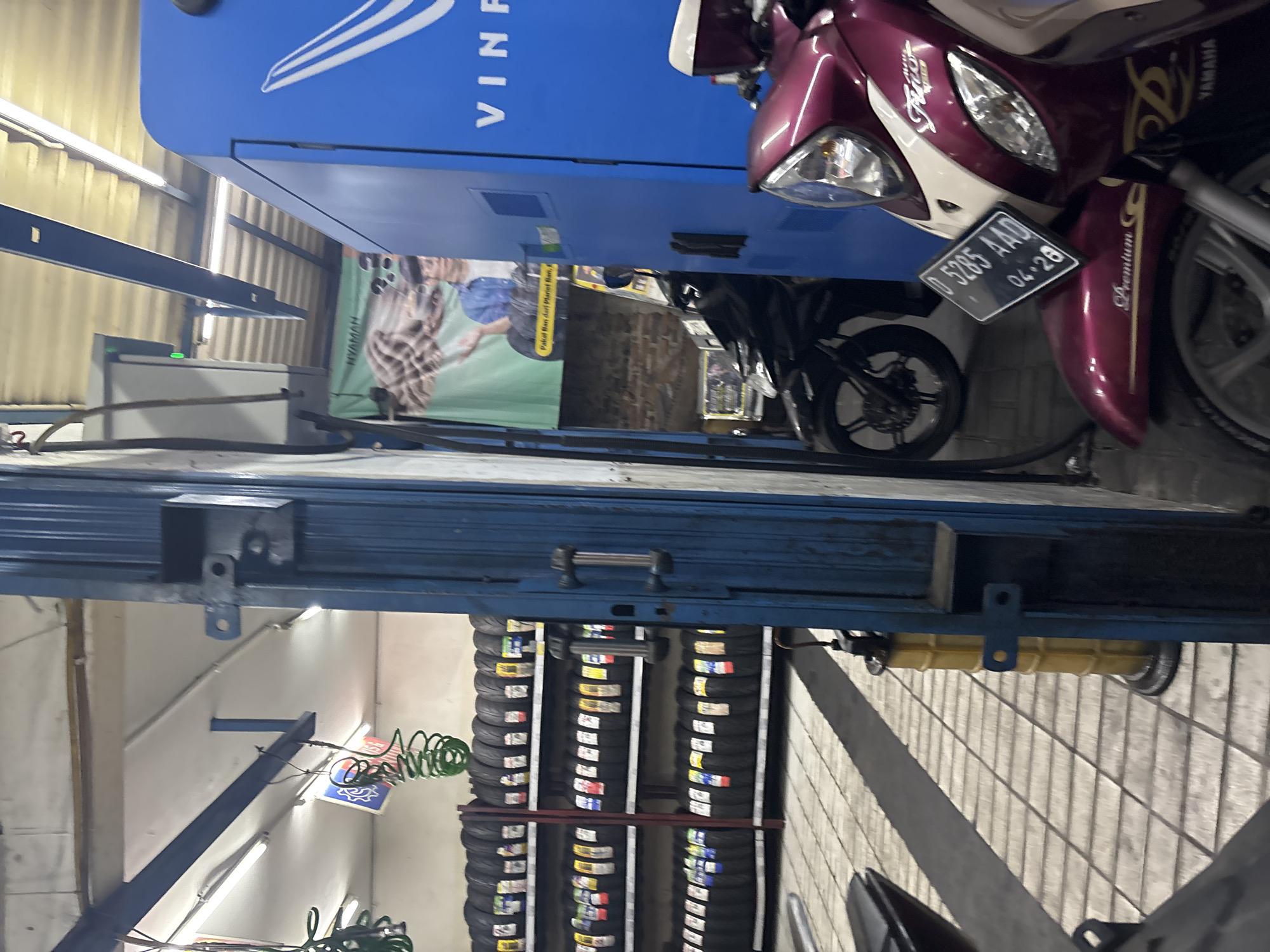 This image captures a scene within a motorcycle repair shop in North Jakarta, Indonesia. The foreground features the front end of a dark red and white Yamaha Fino motorcycle, with its license plate clearly visible: B 5285 AAD, dated 04-28. Behind it, another motorcycle, mostly black, is partially obscured.
Dominating the right side of the frame is a large, bright blue structure, possibly a charging station or storage unit, with the "VINF" logo visible at the top. To the left, shelves are stacked high with rows of new motorcycle tires. A yellow oil drain container stands on the tiled floor, and above, various tools and equipment are suspended from the ceiling, including coiled green hoses.
The background reveals a brick wall and a large green banner with text that includes "NYAMAN" and a depiction of a person. The environment appears to be an indoor, well-lit workspace, typical of a vehicle service center.
