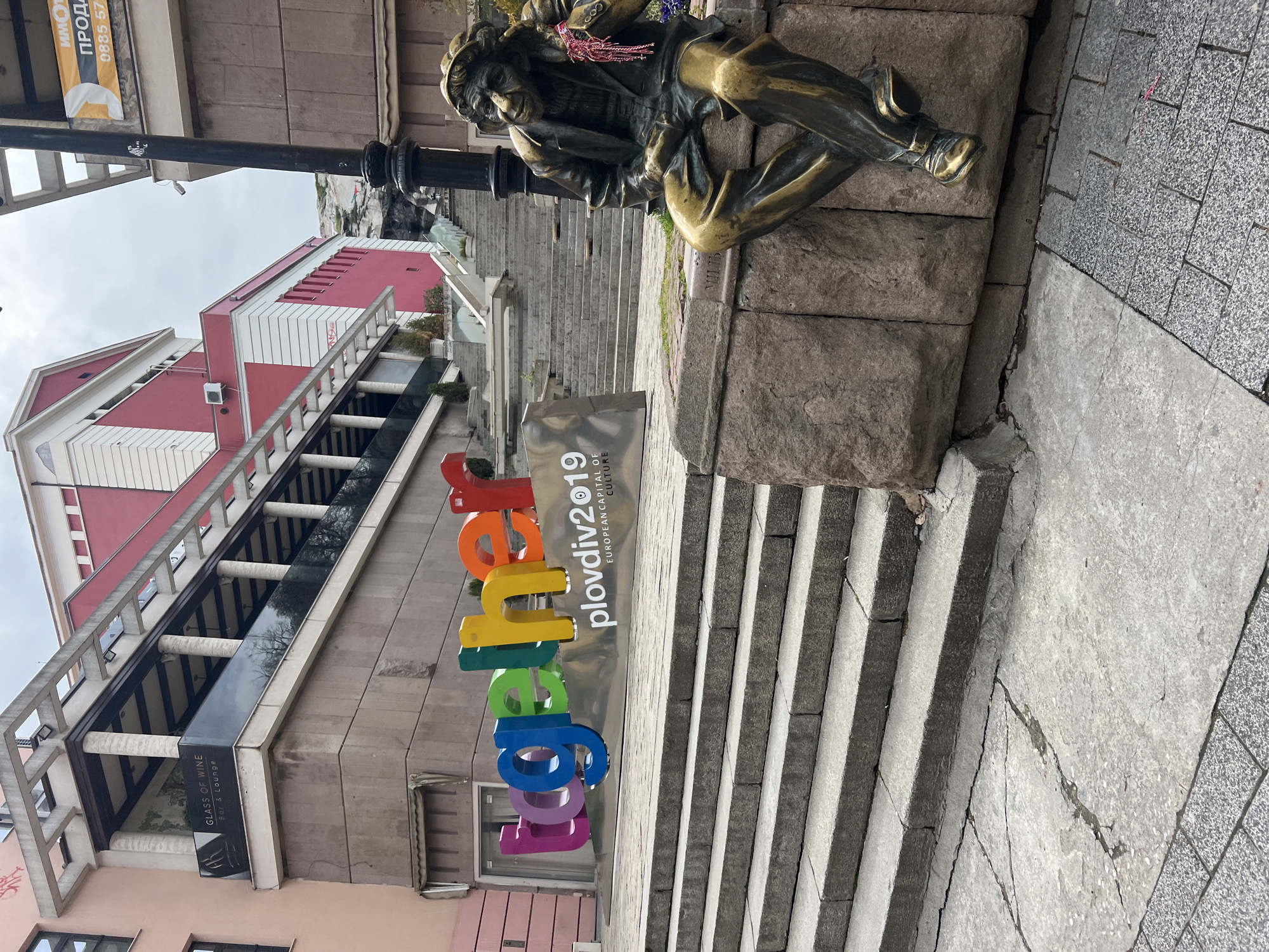 The image captures a scene in Plovdiv, Bulgaria, featuring the "Plovdiv 2019 European Capital of Culture" sign in colorful block letters. The sign stands in front of a set of stone stairs leading upwards. Seated on the stone wall next to the stairs is a bronze statue of a contemplative figure, possibly a writer or artist. In the background, there's a red and white building with a balcony, identified as "Glass of Wine Bar & Lounge" through a sign. A black street lamp is also visible beside the statue. The foreground shows a paved area leading to the stairs and sign. The scene appears to be located in an urban area, presumably a cultural or historical district of Plovdiv.