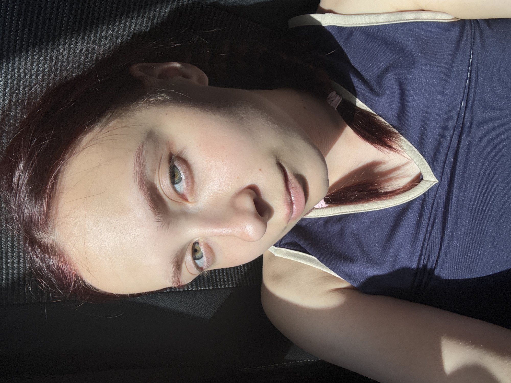 A young woman with light skin and green eyes lies on a dark surface. She has reddish-brown hair and is wearing a dark blue tank top with a light-colored trim. The image is brightly lit in certain areas, creating strong contrast. The specific location in Alnashi, Russia, cannot be determined from this image.