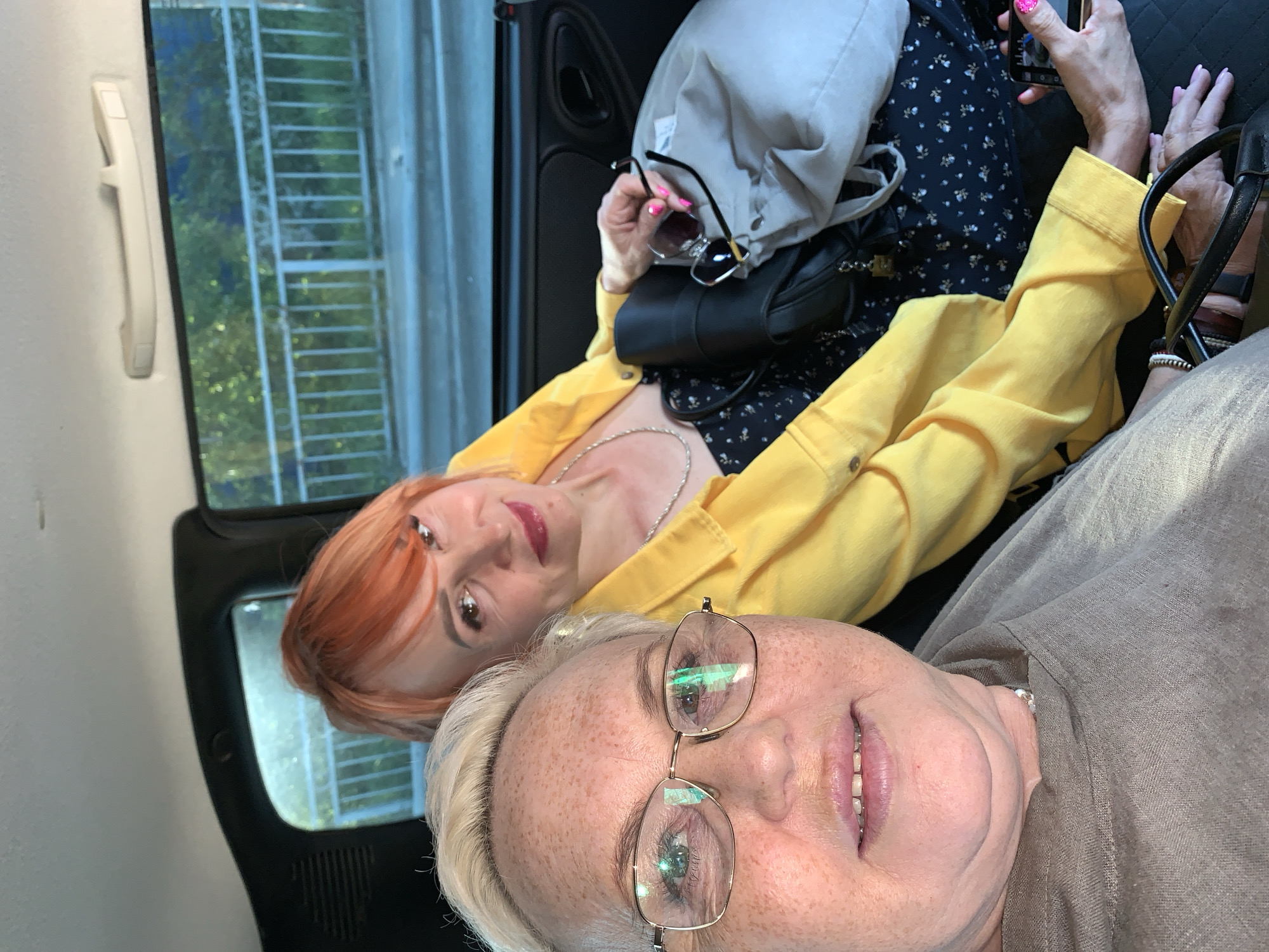 Two women are posing for a selfie in the back of a vehicle. The woman in the foreground has short, blonde hair and is wearing glasses and a grey shirt. The woman in the background has red hair and is wearing a yellow jacket over a dark floral dress. The setting appears to be a car in Chaikovsky, Russia, as indicated by the background seen through the car window, which shows a building with a ladder and a window handle attached to the side of the car. The woman in the background is holding sunglasses and a smartphone. A grey bag and a black purse are visible next to her. The women are looking directly at the camera.