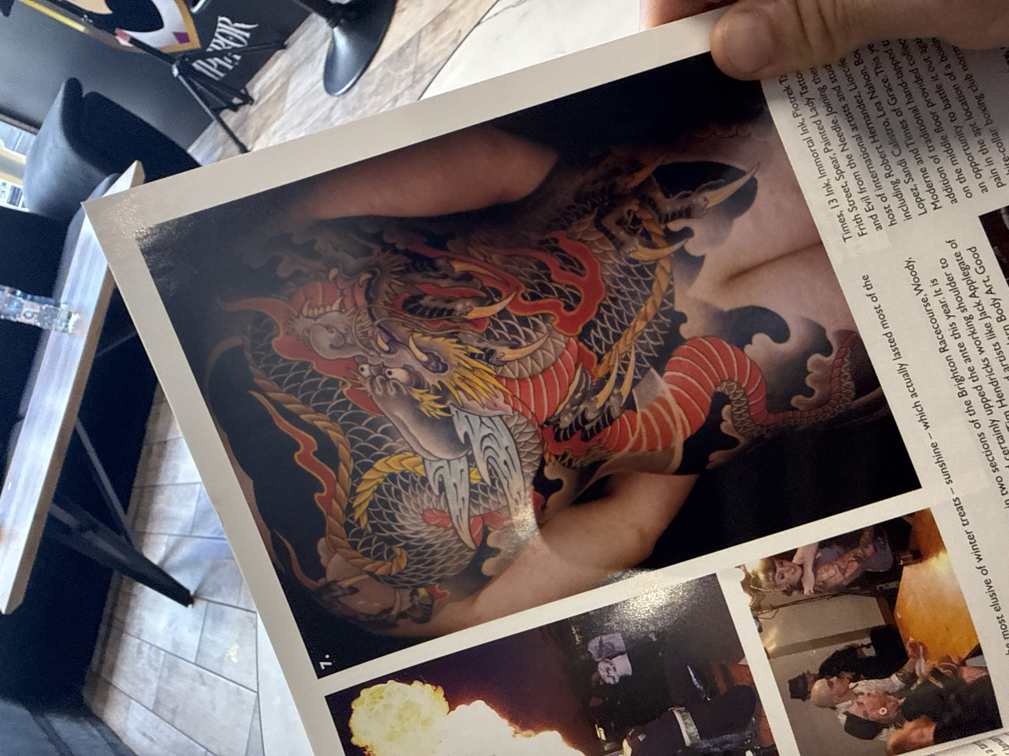 A person is holding a page from a magazine, presumably about tattoos. The magazine page showcases various photographs, including a prominent image of a dragon tattoo covering a large portion of someone's arm and shoulder. The tattoo is detailed, featuring vivid colors like red, gold, and black. The lower portion of the magazine page displays other pictures, one showing a bright explosion, and another depicting a social scene indoors with several people. The scene appears to be in an urban environment and also include barstools. The magazine page also contains printed text about tattoo artists. This image is not in the city of Sofia, Bulgaria.