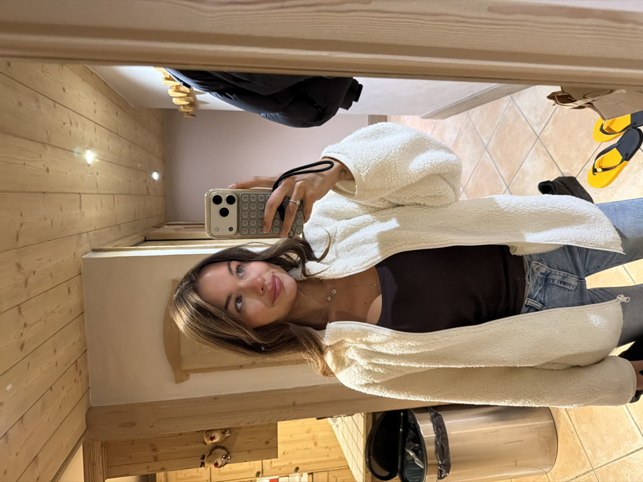 A person wearing a cream-colored fleece jacket, a dark top, and jeans is taking a selfie in front of a mirror. The reflection shows a room with light wood paneling on the walls, a tiled floor, and various objects including a coat, sandals, and a trash can. The person is holding a phone with a light-colored case. The image is slightly rotated.
