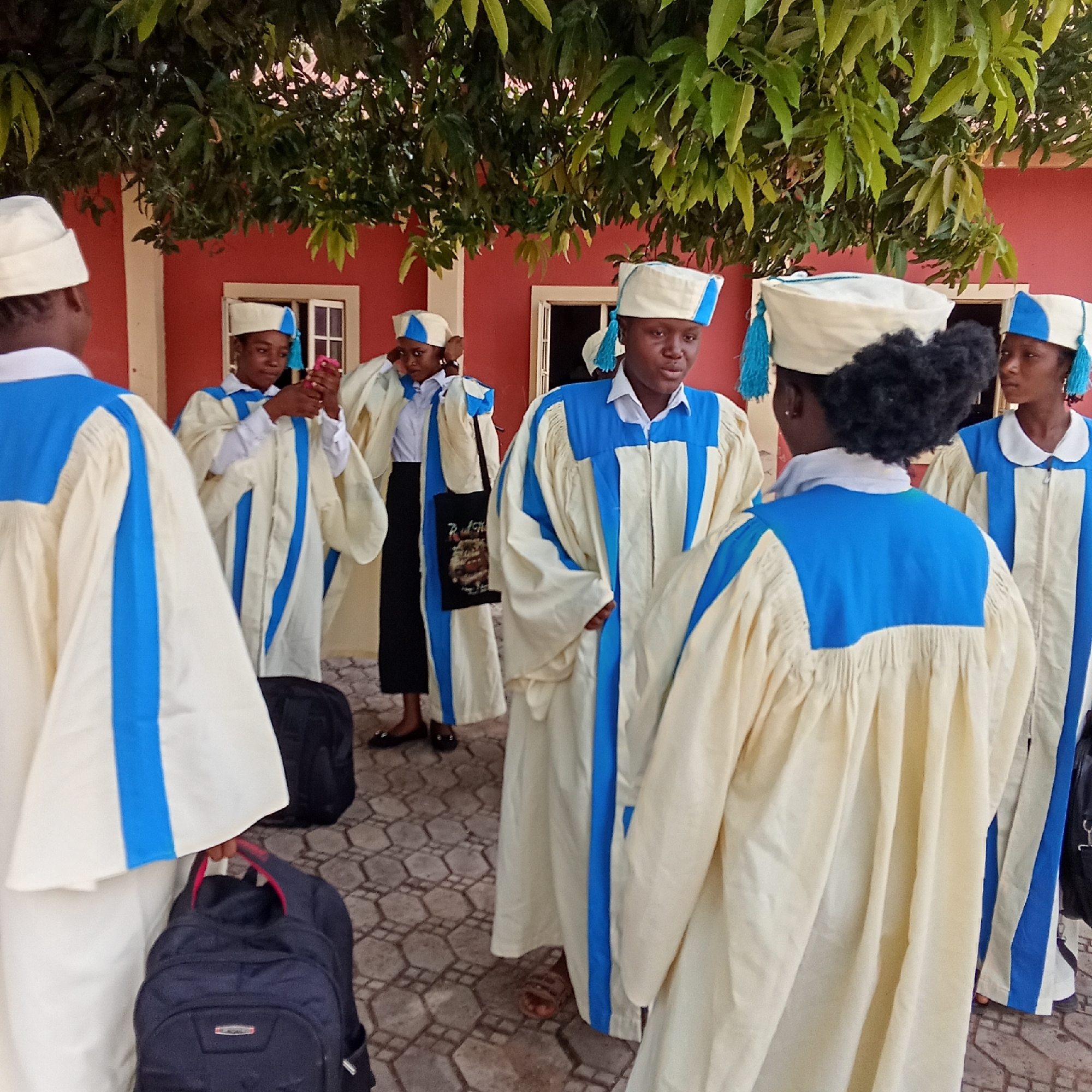 In an outdoor setting, presumably in a city of Unknown, Unknown, a group of individuals are gathered. They are wearing identical light-colored robes adorned with blue accents, along with matching headwear that includes blue tassels. One person is holding a smartphone, possibly taking pictures. Some individuals have bags near them. The backdrop features a red building with a window and a tree providing shade overhead. The ground is covered with a patterned pavement.