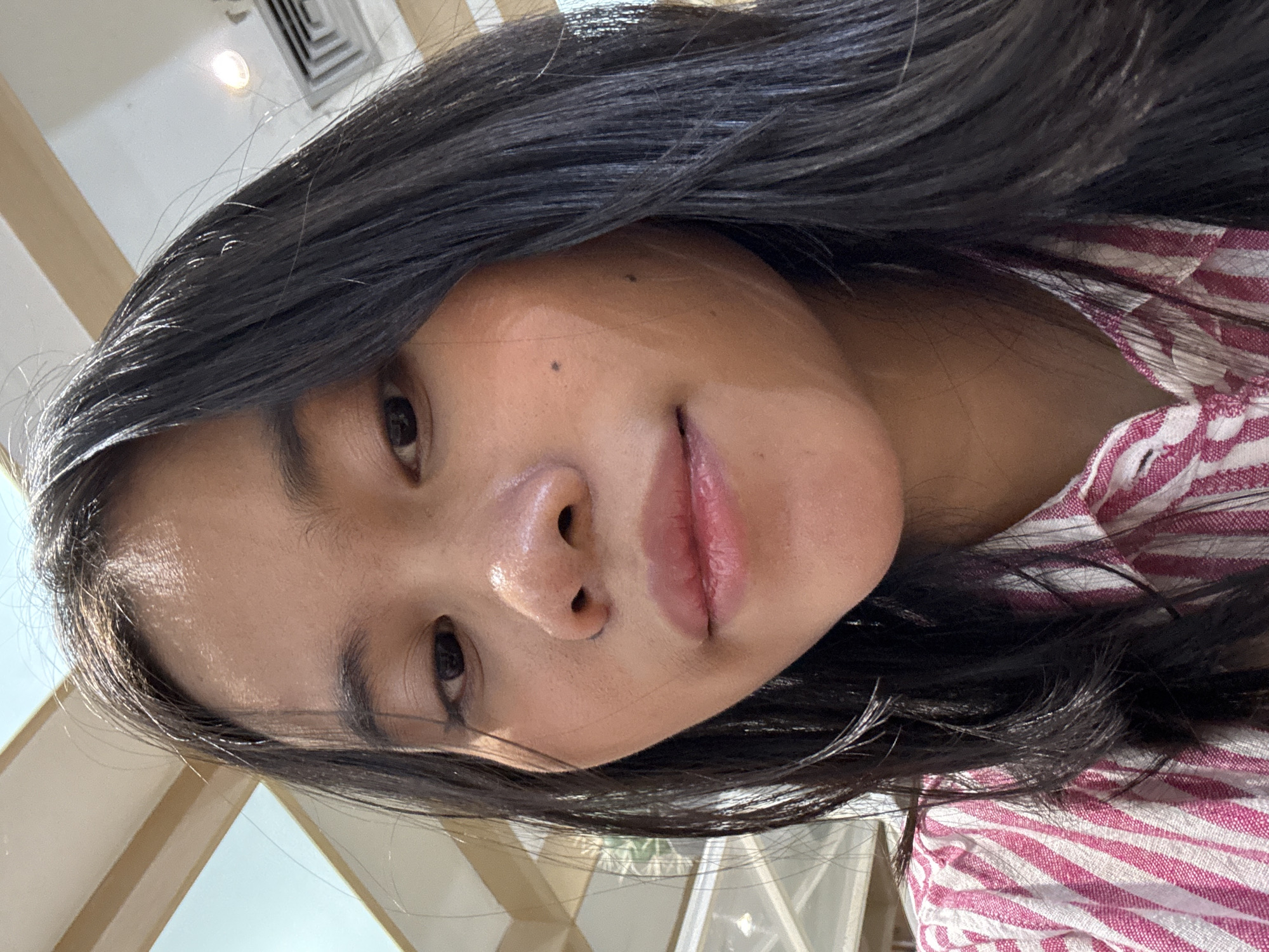 This is a close-up, low-angle, headshot of a young person. The subject has long, dark, straight hair that frames their face. Their skin appears to have a slight sheen, possibly from sweat or a dewy complexion. They have dark eyes, dark eyebrows, and full lips. A small mole is visible on their forehead, and another on their cheek. The person is wearing a pink and white striped shirt.
The background is blurred, but suggests an indoor setting with a fluorescent light fixture and air vents visible in the upper left corner. The lighting is bright, casting a soft glow on the subject's face. The overall impression is casual and informal. The orientation of the image is rotated 90 degrees counter-clockwise. The setting appears to be Central Jakarta, Indonesia.
