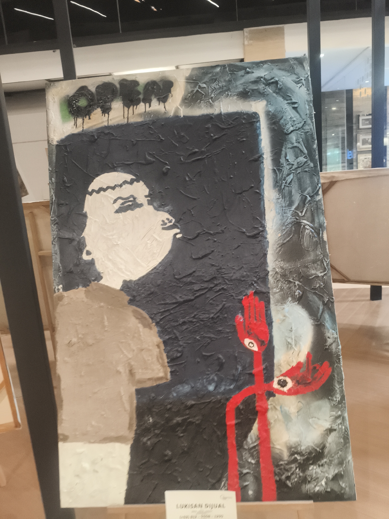 The image displays a vertical, abstract painting with a dark gray textured background. On the left side of the painting, a stylized face with white skin and closed eyes is depicted in profile, looking to the right. The face has a zigzag pattern for hair and a beige-colored garment underneath. To the right of the face, a bright red, stick-figure-like character with two eyes on its "head" is shown. This figure has two outstretched hands, each with a single eye in the palm. The painting is displayed on a stand in what appears to be an indoor exhibition space, possibly a gallery or a shopping mall, given the presence of reflective surfaces and shelving in the background. A small white plaque with text is visible at the bottom of the painting, suggesting it is for sale or exhibition. The overall impression is one of contemporary art with a somewhat unsettling or thought-provoking theme. The lighting is even, indicating indoor ambient light.