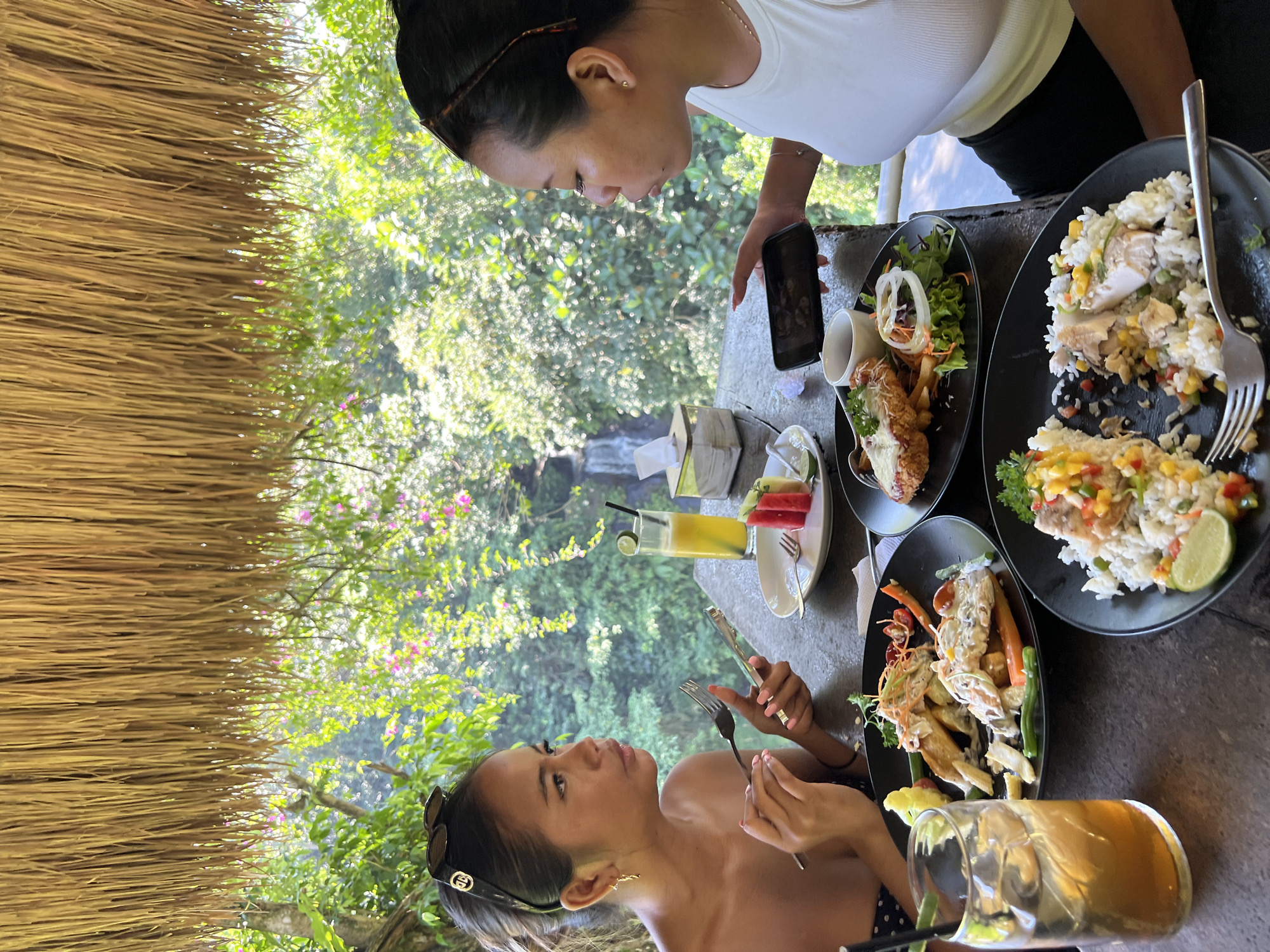 Two young women are seated at a table with food and drinks, outdoors, in what appears to be a restaurant with a thatched roof. The background reveals a lush green forest with a visible waterfall. The woman on the left, wearing a dark top with white polka dots and sunglasses on her head, holds a fork in her left hand and a smartphone in her right. She is looking to her left. The woman on the right, wearing a white tank top, is looking down at her smartphone, which she holds in her left hand. On the table are two black plates with food, one containing rice with colorful vegetables and a piece of fish, the other with a breaded cutlet, fries, and salad. A glass of amber-colored iced tea with a straw and lime wedge is also on the table, as is a small plate with watermelon cubes and lime. A tissue dispenser is visible behind the woman on the left. The overall setting suggests a tropical location in Blahbatuh, Indonesia.