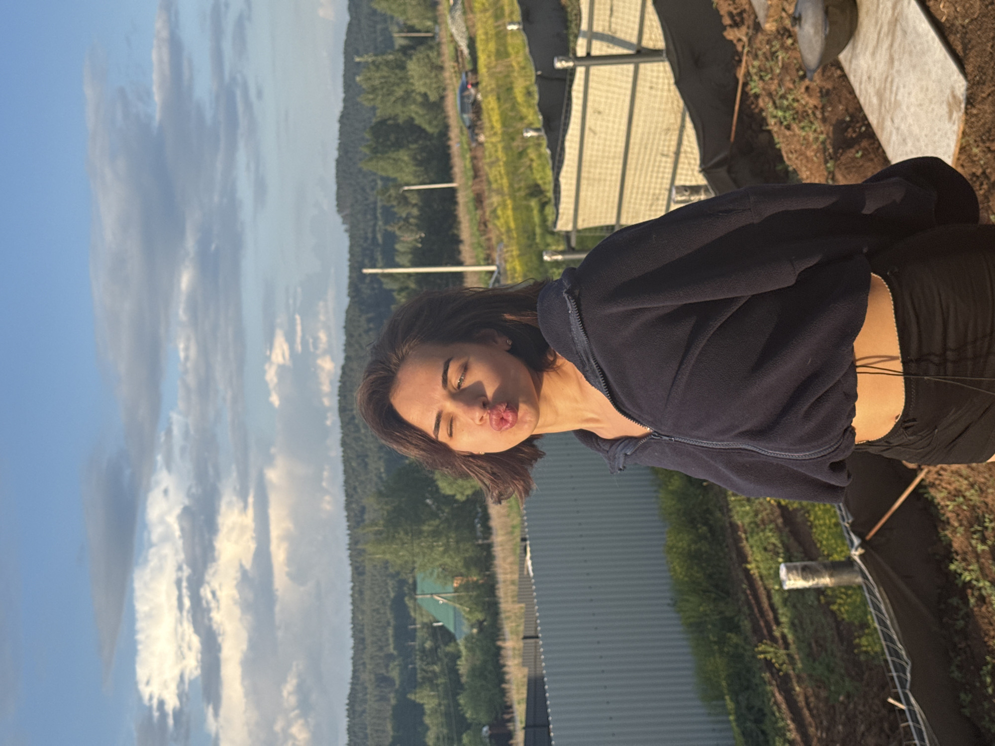 A woman is posing for a photo outdoors. She is puckering her lips and has her eyes closed. She is wearing a dark-colored zip-up jacket and dark shorts. The background features a landscape with trees and a cloudy sky. Some metal structures are also visible. This photo was taken in Лобаново, Russia.