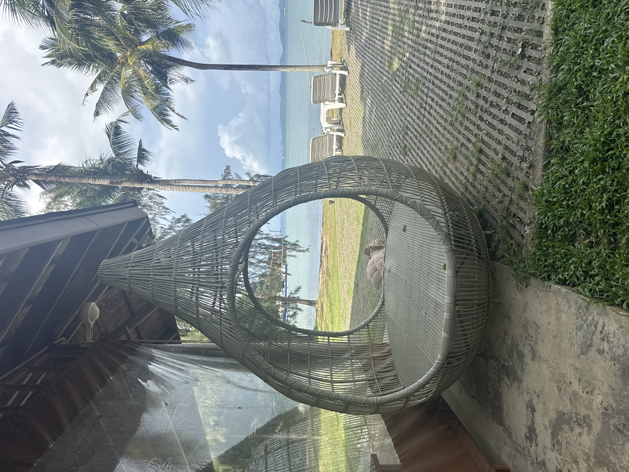 This image depicts an outdoor scene at a resort in Pa Khlok, Thailand. In the foreground, a large, teardrop-shaped woven hanging chair is prominently featured, positioned on a concrete patio. The chair appears empty. Behind the chair, a lawn extends towards a body of water, likely the Andaman Sea. Several lounge chairs and a small table are visible on the lawn, facing the water. Coconut palm trees line the background, and the sky is partly cloudy. The overall impression is one of a tranquil, tropical vacation setting.