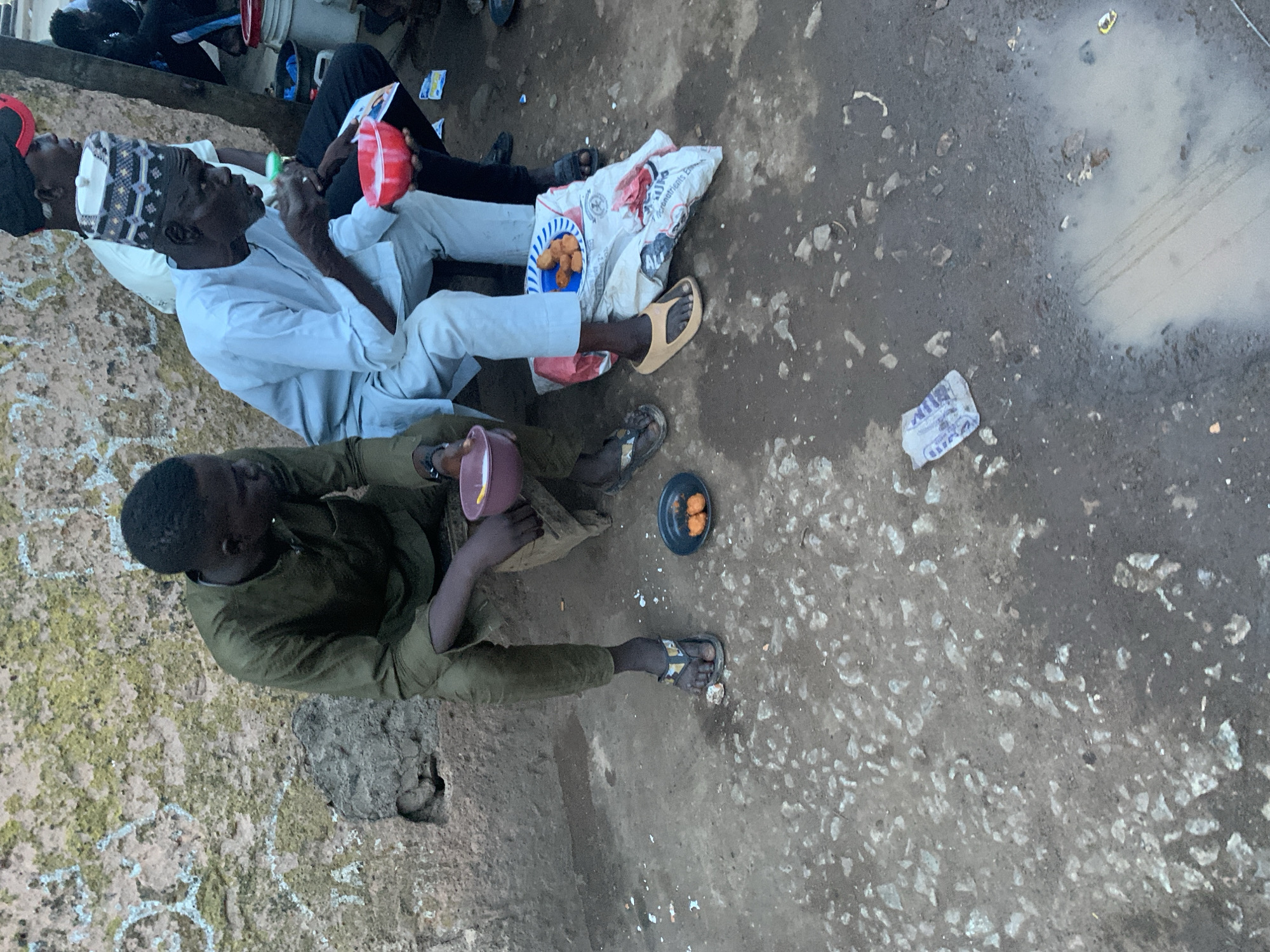 The image, taken in Gura Top, Nigeria, shows two men and other partially visible individuals in an outdoor, informal setting during the daytime. The ground is composed of rough dirt and small stones, with noticeable patches of wet mud and puddles, indicating recent moisture.
In the midground, a man wearing a light-colored traditional outfit and a patterned cap sits, holding a red plastic bowl. Beside him, on a white sack or bag, a blue plate holds several golden-brown, fried dough-like items. He wears light brown slip-on sandals.
To his right, another man, dressed in a dark green or olive-colored top and trousers, sits on the ground. He holds a purple-brown plastic bowl. Near his feet, a small black plate contains two round, yellow items, likely boiled eggs or a similar snack. He is wearing patterned flip-flops.
Other people are partially seen in the background to the left. The activity appears to be a casual meal or gathering. The scene suggests a relaxed atmosphere in an everyday, rustic environment.
Visible text includes the word "BIM" on a small piece of paper on the ground. Faint, mostly illegible text, possibly related to "NUTRITION," can be seen on the white sack.