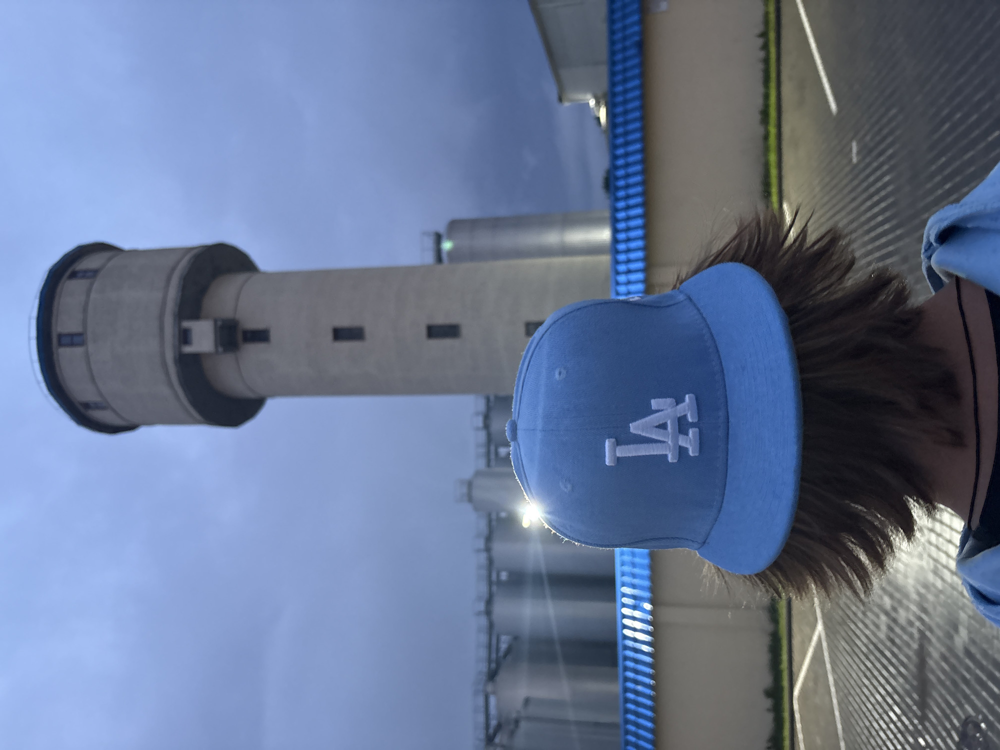 A person wearing a light blue baseball cap with a white "LA" logo is shown from the back, with their brown hair visible. The cap is tilted, with the brim catching a bright light that creates a lens flare effect. The person is standing in an outdoor setting at dusk, indicated by the dim, cool-toned lighting and the sky's appearance.
In the background, a tall, cylindrical concrete structure, possibly a water tower or industrial silo, dominates the left side of the image. It has several rectangular window-like openings and a dark band around its top. To the right of this structure are several more, smaller cylindrical silos or tanks, also appearing to be industrial in nature. These are positioned in front of a light-colored building or wall. A bright blue, slatted structure runs vertically along the right side of the background, partially obscuring the building behind it. The ground in the foreground appears to be a paved surface with a distinct striped shadow pattern, suggesting the presence of a fence or grate.
The overall mood is somewhat subdued and observational, with the focus on the person and the industrial backdrop. The lighting suggests it is either early morning or late evening. There is no discernible weather, but the sky is overcast or dusky. The image was taken in Lida, Belarus.