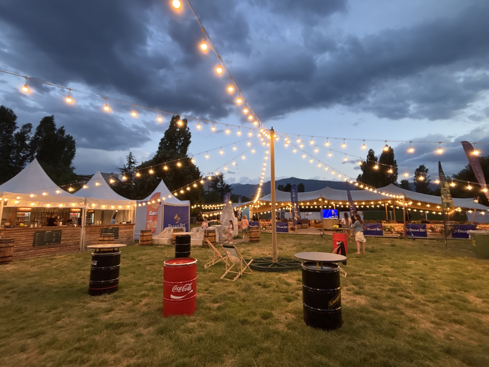 This image depicts an outdoor event space at dusk in Sofia, Bulgaria, illuminated by strings of warm, glowing lights. The scene is set against a backdrop of a cloudy, twilight sky with hints of mountains in the distance.
In the foreground, a grassy field is dotted with several repurposed barrel tables. One prominent table is a red Coca-Cola barrel, another is a black barrel with "Schweppes" branding, and a third is a black barrel with a more generic design. Scattered throughout the area are lounge chairs.
Dominating the middle ground are several large, white tents, likely serving as booths or bars. One tent on the left appears to be a functional bar with shelves stocked with bottles and a person visible behind the counter. Another tent further back has banners displaying the text "PALMS BET". People are visible milling around the tents and sitting in the chairs, suggesting an active event.
The overall ambiance is festive and relaxed, characteristic of an outdoor gathering or festival. The string lights create a warm and inviting atmosphere as the natural light fades.