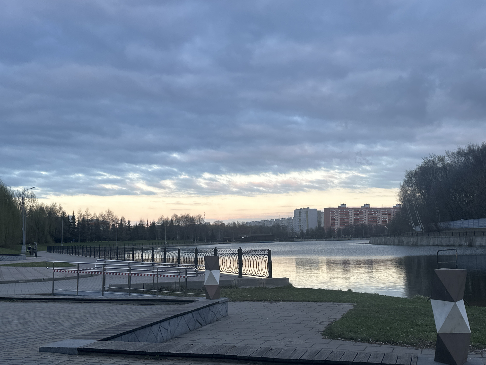 The image depicts a park scene in Moscow, Russia, likely near the Golyanovo Pond. The sky is heavily overcast with grey clouds. A body of water, presumably the pond, stretches across the midground, reflecting the sky's pale light. A black iron fence runs along the edge of the water.
In the foreground is a paved walkway with a ramp and stainless steel railing, partially blocked off with red and white tape. A modern, geometrically designed trash receptacle stands nearby.
Across the pond, a line of trees is visible, interspersed with residential apartment buildings. Two figures can be seen walking along the promenade on the left side of the image. The overall impression is of a quiet, overcast evening in a park setting.