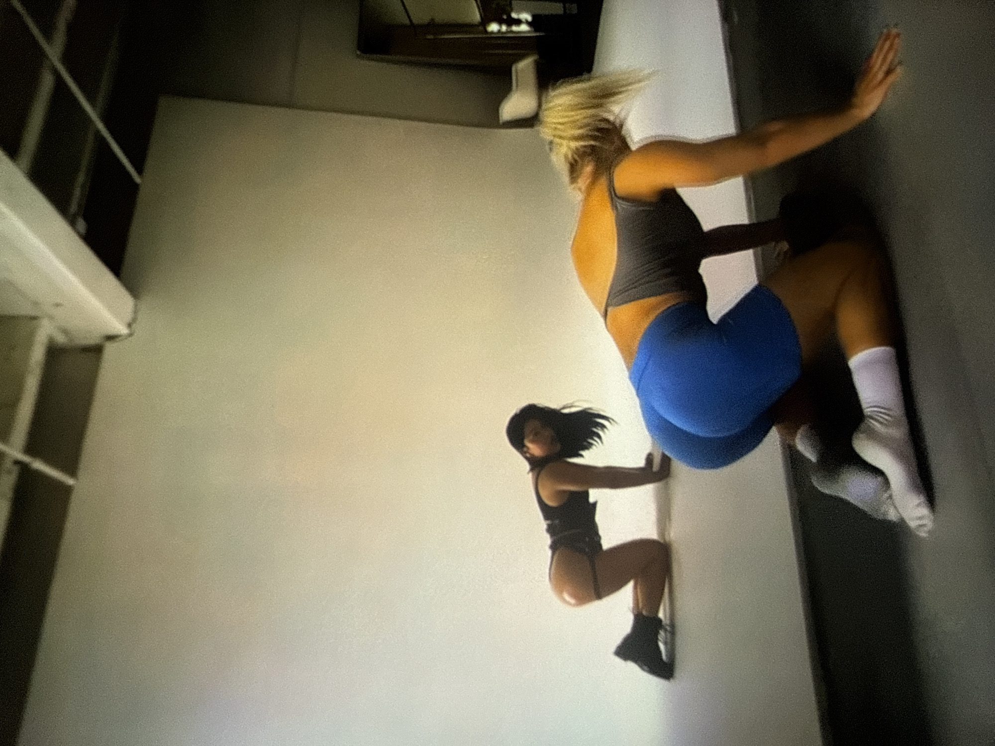 The image displays an indoor scene, identified as being in Varna, Bulgaria, featuring two individuals positioned on a dark grey floor against a plain, light-colored seamless backdrop wall. The ceiling is dark with visible white structural beams and a white cable. A large, dark, angled reflective surface, possibly a mirror, is present on the right side of the background, with a partially visible light-colored chair-like object.
Two individuals, both appearing female, are positioned on their hands and knees. The individual positioned further back on the left has dark hair, wears a dark sleeveless top or bodysuit, and dark boots. A dark strap is visible around her right upper thigh/buttock. Her face is partially visible as she turns her head slightly towards the viewer.
The individual in the foreground on the right has blonde hair tied in a high ponytail. She is wearing a dark grey sports bra and bright blue shorts, along with white socks. Her head is directed downwards, and her back is arched, elevating her buttocks.
Both subjects are in a similar all-fours pose, facing away from each other. The lighting is diffused, and the image exhibits visible digital noise or grain.