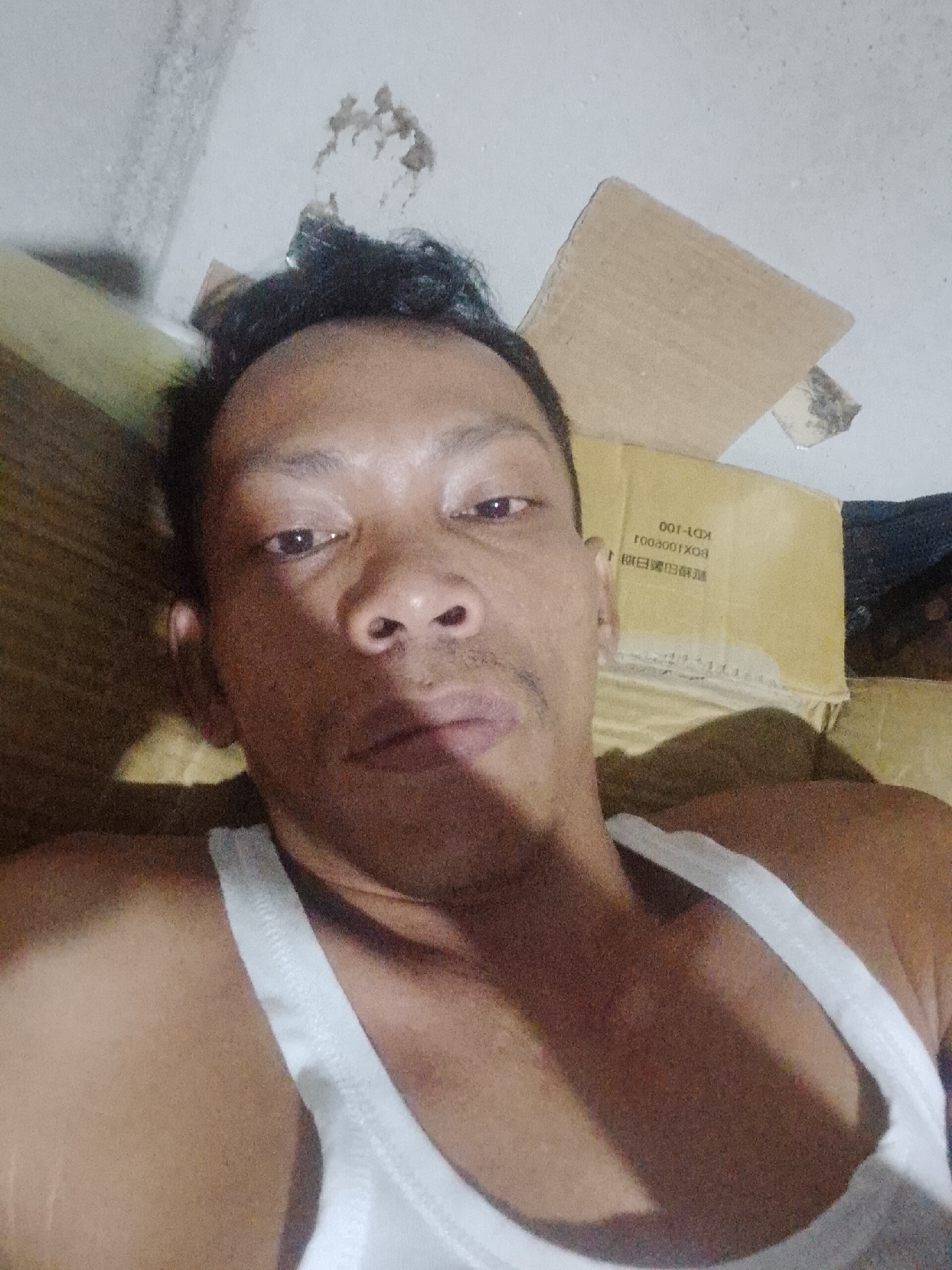 A medium close-up shot features a man wearing a white tank top. He is indoors, lying down near cardboard boxes. The location is Bukit Ketok, Indonesia. The wall behind him shows signs of discoloration or damage.