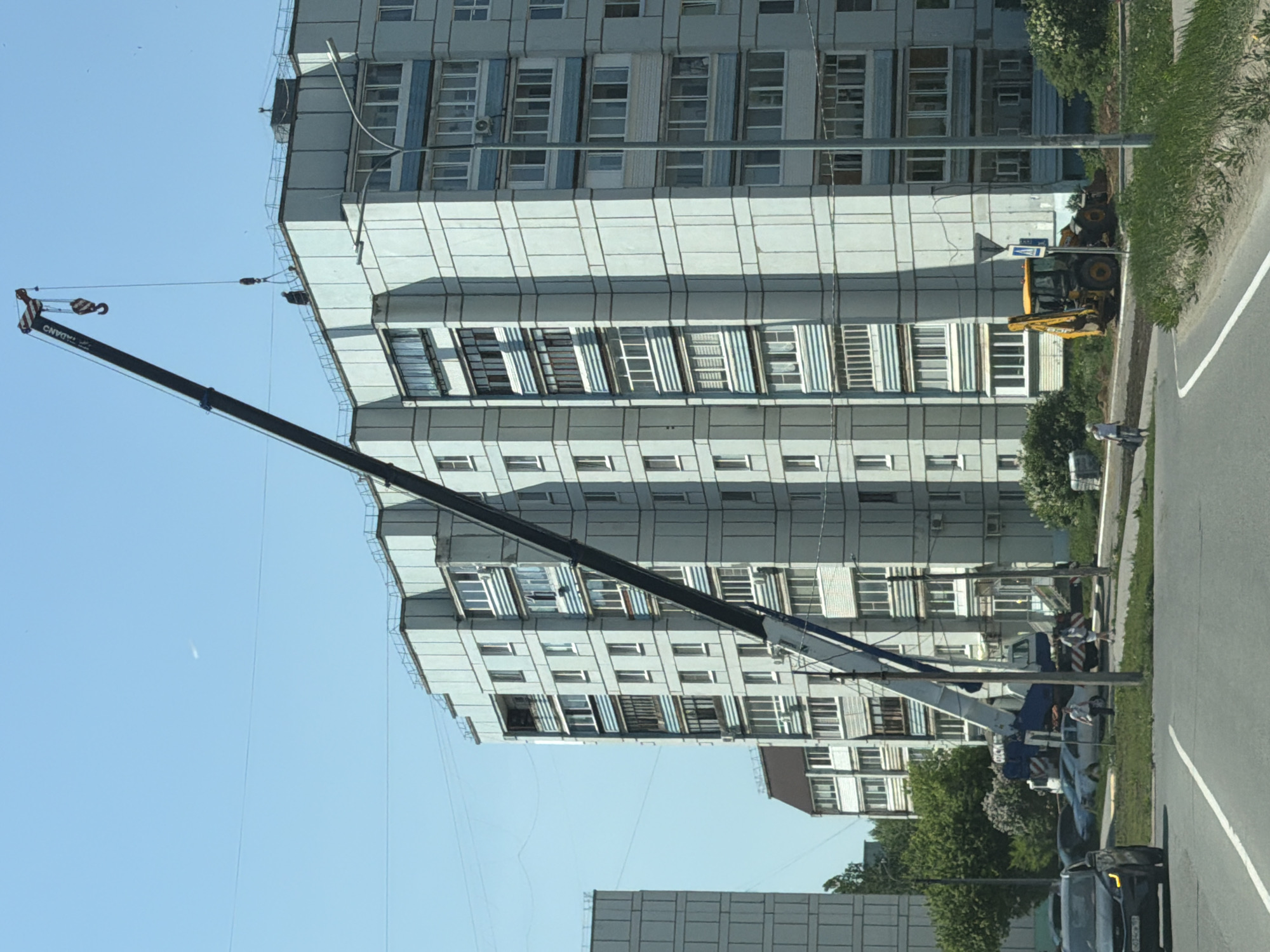 The image shows a mobile crane with its boom extended towards a multi-story apartment building. The crane, displaying the text "TADANO" on its boom, is positioned on the side of a road in a built-up area. A construction vehicle, possibly a backhoe loader, is parked near the building. A person is walking on the sidewalk near the construction equipment. The backdrop consists of residential buildings, trees, and a clear blue sky. Based on the architectural style of the buildings and the presence of specific vehicles, the location is possibly in the city of Chaikovsky, Russia.
