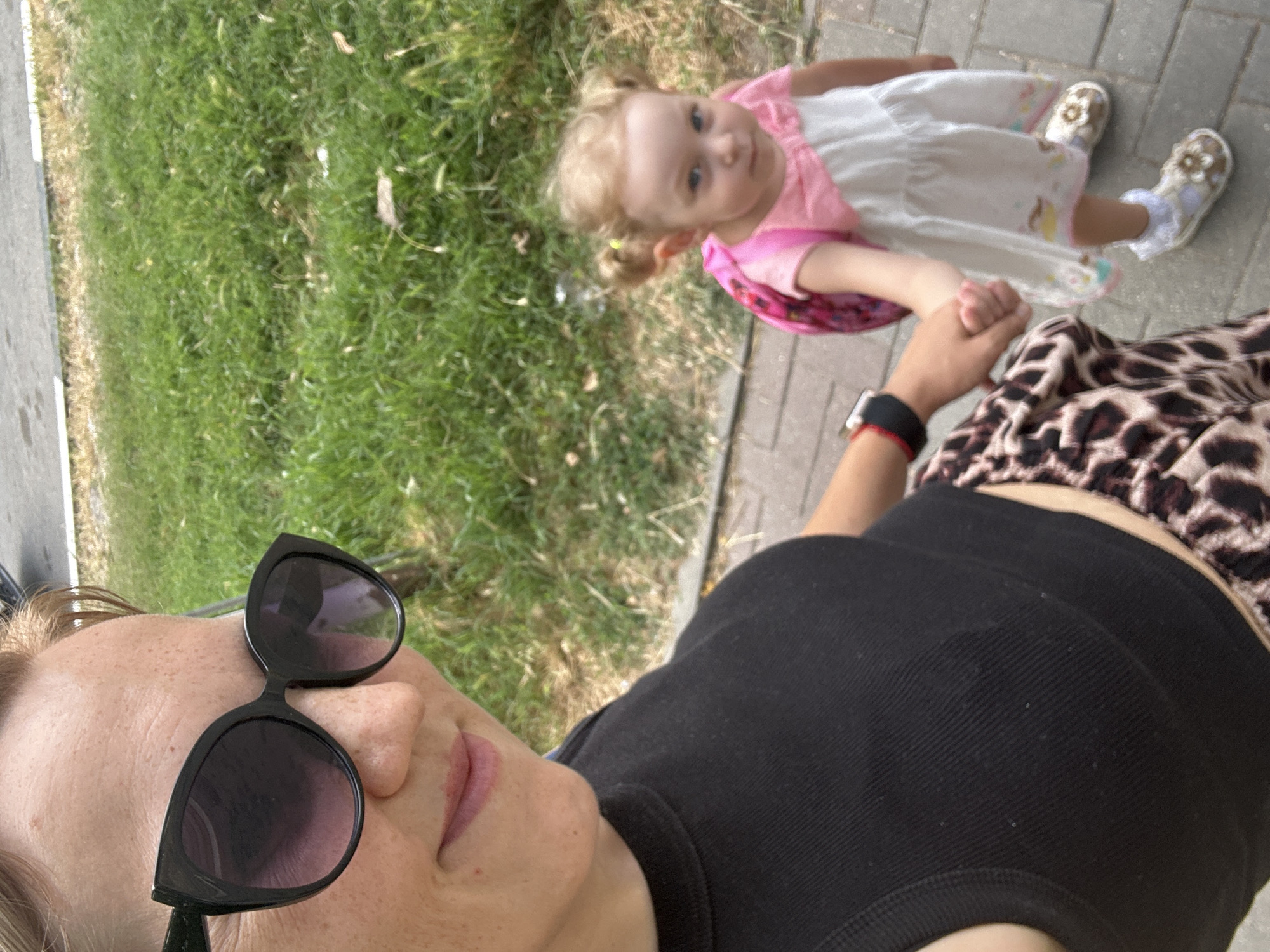 A woman wearing large black sunglasses, a black tank top, and leopard-print pants holds hands with a young child. The child, with blonde hair in two pigtails, wears a pink and white dress and a pink backpack. They are walking on a paved sidewalk bordered by grass. The scene appears to be in Gelendzhik, Russia, indicated by the visible greenery and pavement typical of the city.