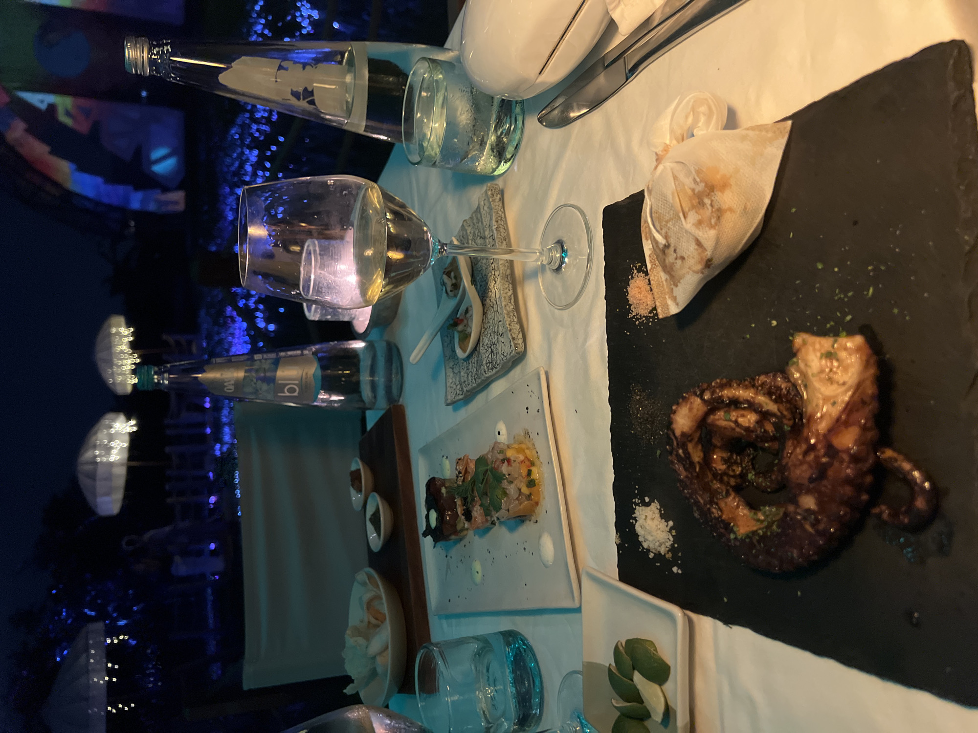 The image displays a well-appointed dinner table at night, likely in an outdoor or open-air restaurant setting in North Kuta, Indonesia. The scene is lit by soft ambient light on the table, contrasting with a dark background featuring blurred blue decorative lights, suggesting an evening dining atmosphere.
The table is covered with a white tablecloth and features various dishes and drinks. A prominent dish is a large portion of grilled octopus, artfully arranged on a dark slate plate with a sprinkle of herbs and coarse salt. Beside it on the same plate is a small, light-colored cloth pouch, possibly containing bread, with a scattering of pinkish salt. Further along the table, a rectangular white plate holds a vibrant dish that appears to be ceviche or tartare, garnished with fresh herbs and small drops of sauce. Adjacent to this, a small wooden board supports two tiny white bowls containing sauces or condiments.
Beverages include two clear glass water bottles, one visibly labeled with "bl" (likely "blue"). Two wine glasses are present, one containing white wine, alongside two tumblers, one holding water. A small white bowl near the bottom contains light-colored items, possibly bread or crackers. In the lower right corner, a small white dish holds several lime wedges. Cutlery, including a knife and fork, is neatly arranged on the tablecloth. The overall presentation suggests a refined and upscale dining experience.