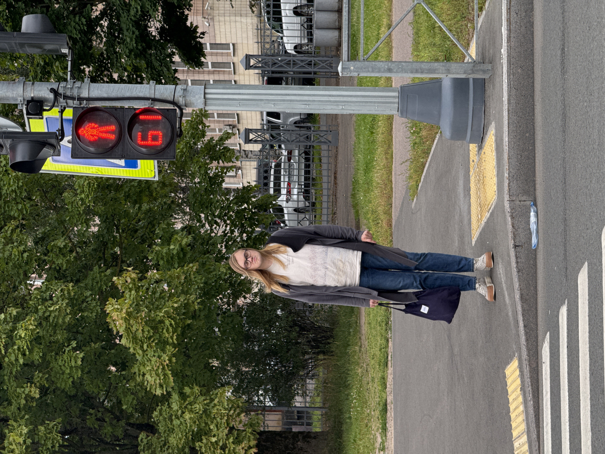 A person stands on a sidewalk at a crosswalk in Sinyavino, Russia. The person is wearing glasses, a light-colored t-shirt with a graphic print, blue jeans, a long gray cardigan, and light-colored sneakers. They are holding a dark blue tote bag in their left hand. To the right of the person is a traffic light pole displaying a red walking signal and a countdown timer showing "61". In the background, there are trees, a building, and parked cars behind a fence. The sidewalk has tactile paving and a raised curb. A plastic bottle is visible on the ground near the curb. The overall lighting suggests an overcast day.