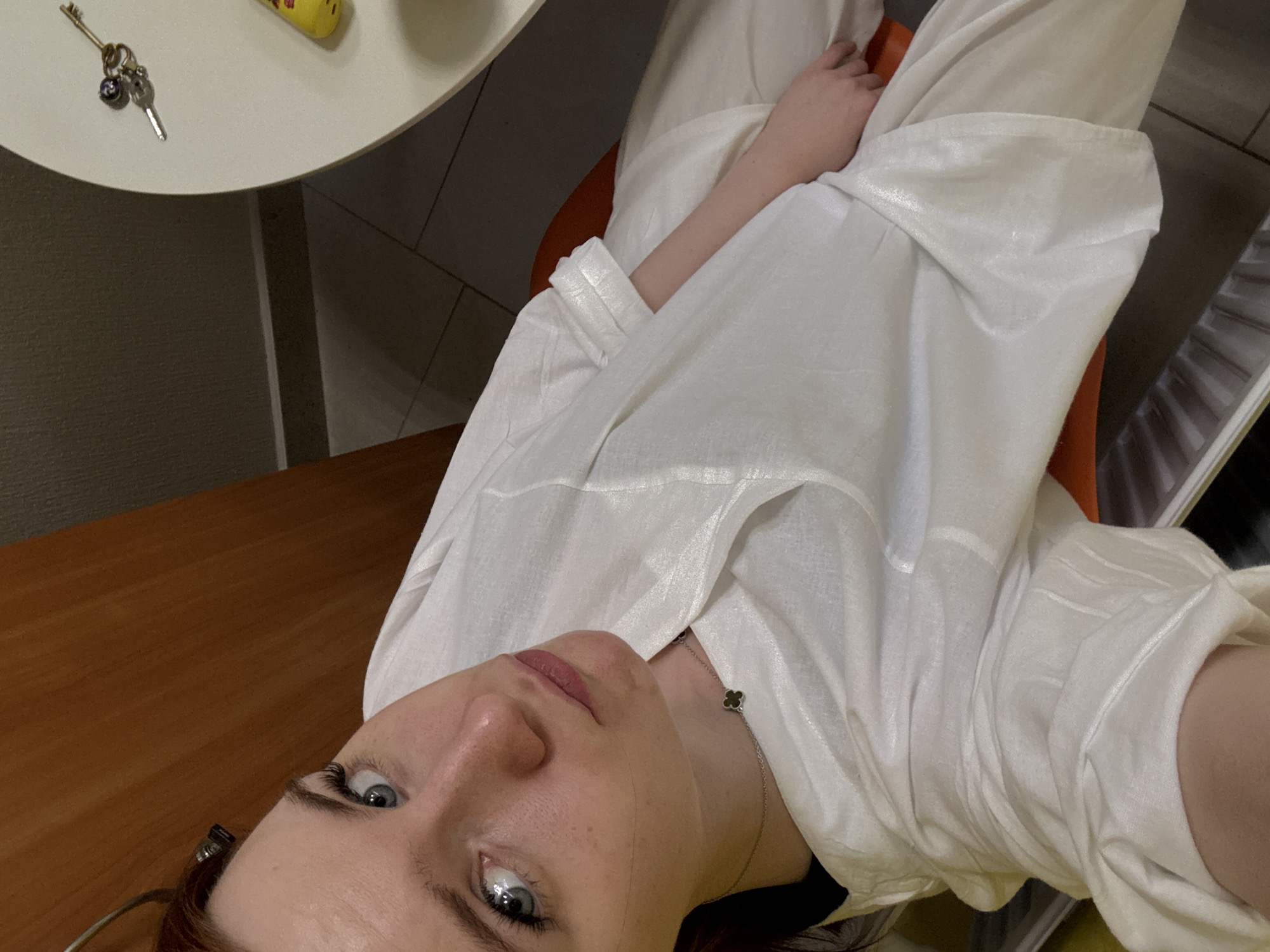 A woman with light skin and blue eyes is looking up at the camera. She is wearing a white, loose-fitting shirt and a delicate necklace. A small round table with keys and a yellow object rests to her left. A wooden desk is visible below. The location is not discernible from the image.