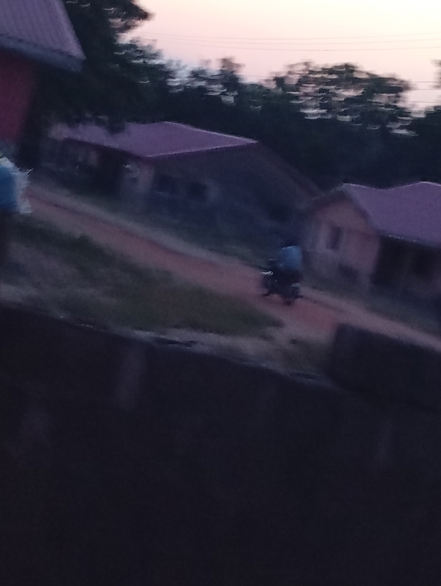 The image shows a dirt road running through what appears to be a residential area in a rural location. Several small buildings with red roofs can be seen along the road. A single person is riding a motorcycle along the road. The environment appears to be tropical with trees visible in the background. The time of day is either dusk or dawn, indicated by the dim lighting and sky color. The specific location within the city of Unknown, Unknown is difficult to determine with the given information.