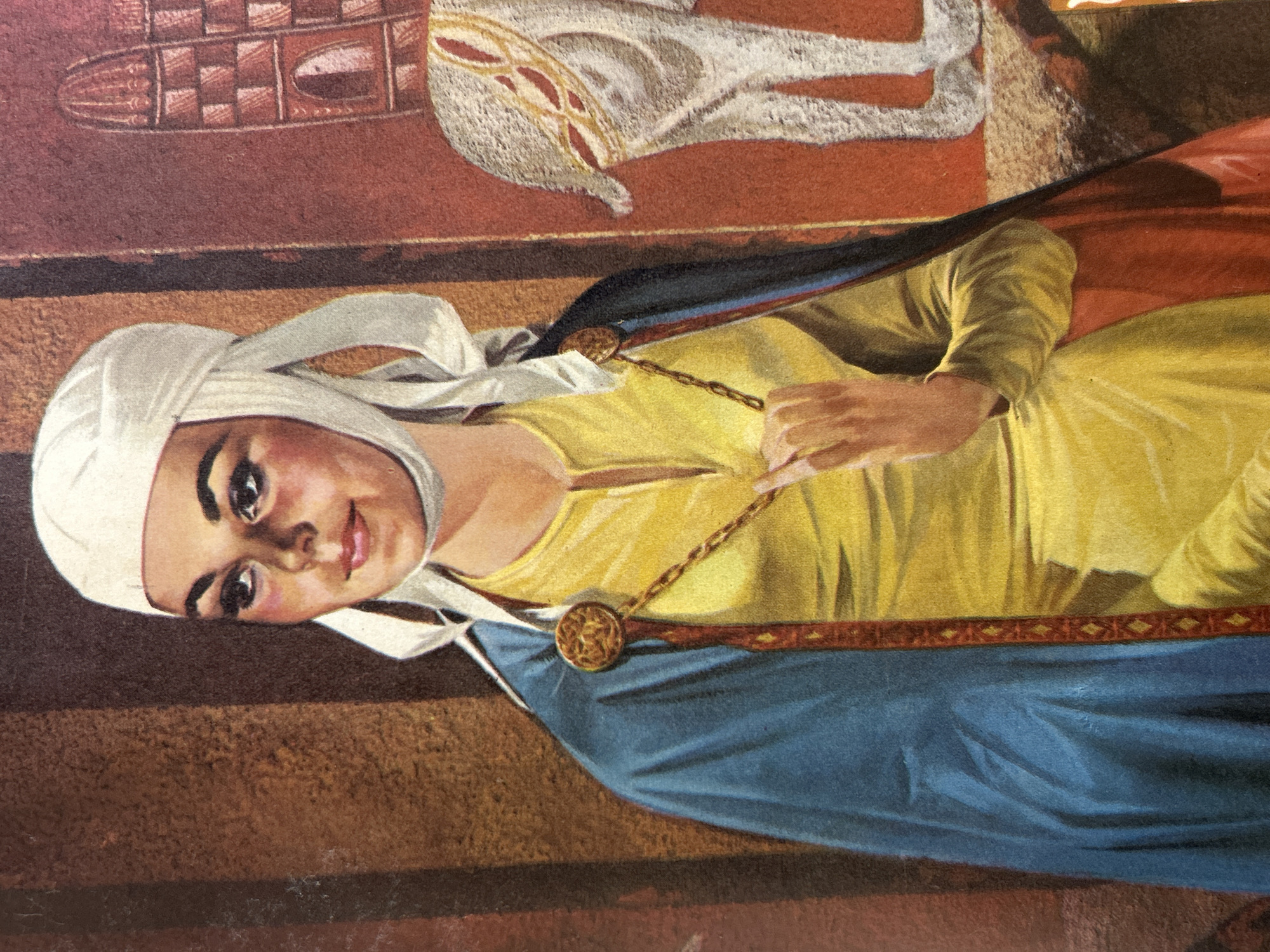 The image is a painting or illustration depicting a young woman wearing a white head covering, a yellow tunic, and a blue cloak. She has dark hair and is looking slightly to her left with a gentle smile. A gold chain with a medallion is visible around her neck, and the cloak is fastened with a circular gold clasp on her left shoulder. The background is composed of textured walls in brown and red tones, with a partial view of a stylized building on the right and what appears to be the hindquarters of a white animal with red and gold decorative elements. The lighting suggests an indoor setting. There is no identifiable information within the image itself that places it in Santa Ana, Venezuela.