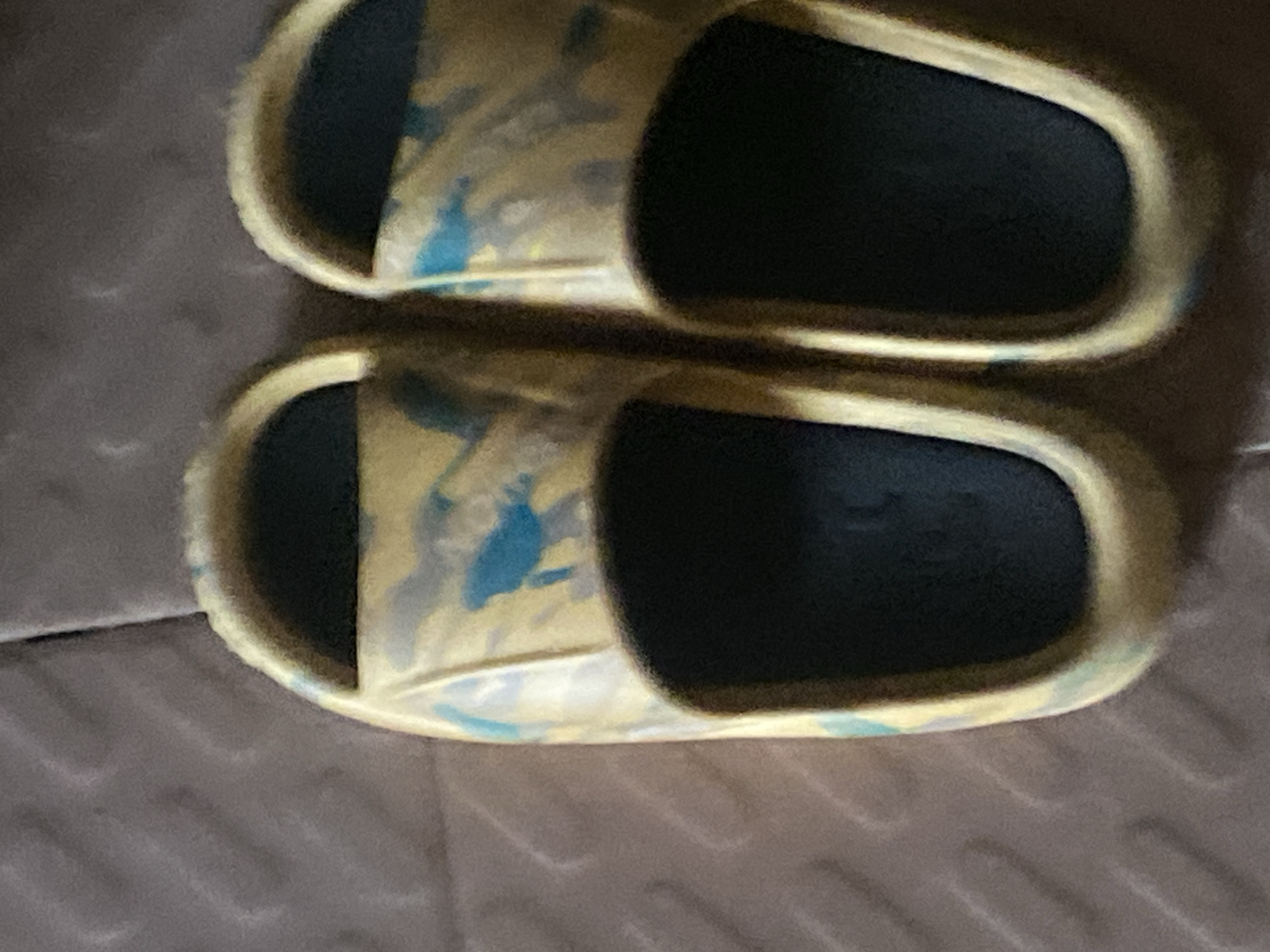 Two yellow and blue camouflage slides are resting on a textured brown surface, likely a floor or mat. The slides are positioned side-by-side, with the left slide slightly in front of the right. The slide on the left appears to have a small tear or imperfection on its left strap. The background is a blurry, muted brown, suggesting an indoor setting. There is no indication of the city of Jauro Yinu, Nigeria, in the image.