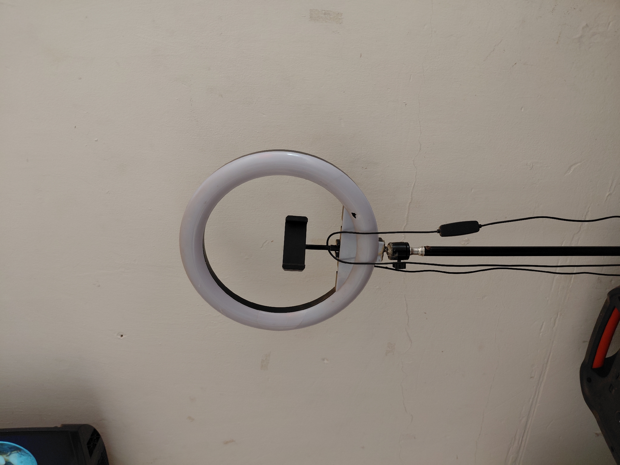The image displays a ring light setup, positioned against a light-colored, textured wall or ceiling. The main subject is a white, circular ring light, appearing to be off, with a black phone holder mounted in its center. Extending from the right side of the ring light is a black metallic pole or stand, which supports the light.
Several black electrical wires are connected to the ring light and run along the pole, with a rectangular black control switch visible on one of the wires, positioned to the right of the ring light. The wall itself is off-white or light beige, showing various imperfections such as subtle cracks, smudges, and areas of discoloration, suggesting an aged or well-used surface.
In the bottom left corner, a portion of a dark, possibly electronic device (e.g., a speaker or monitor base) is partially visible. Similarly, in the bottom right, a part of another dark object with a prominent orange handle is seen. No people are present in the image, and there is no visible text. The setup suggests equipment used for photography, video recording, or live streaming.