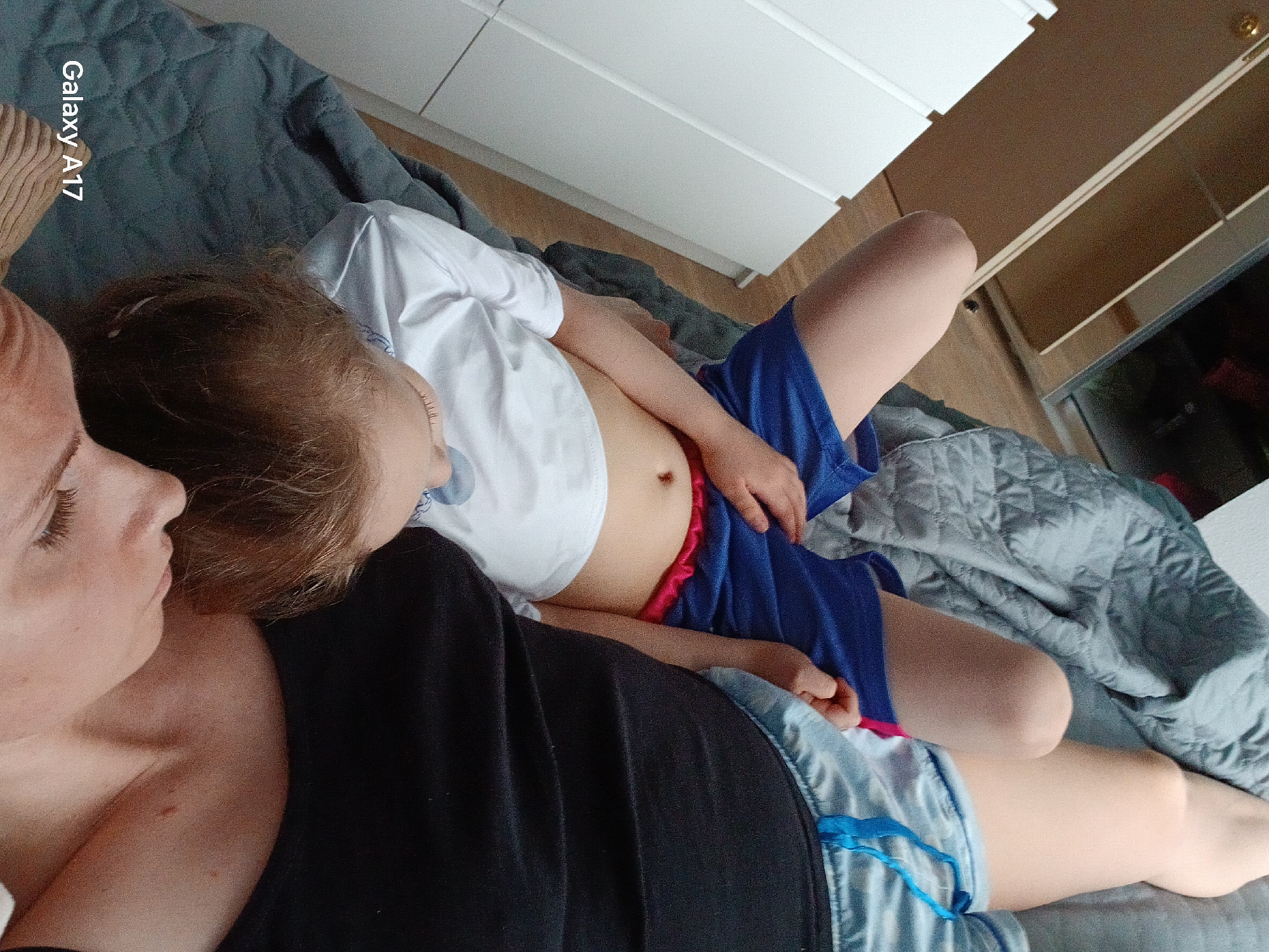 The image shows a woman and a child lying on a bed. The woman is wearing a black top and denim shorts. The child is wearing a white t-shirt and blue shorts with a red waistband. They are both lying on their sides, facing each other. The bed is covered with a gray quilted blanket. In the background, there is a white dresser and a partial view of an interior. There is no information to indicate a location in Yekaterinburg, Russia.