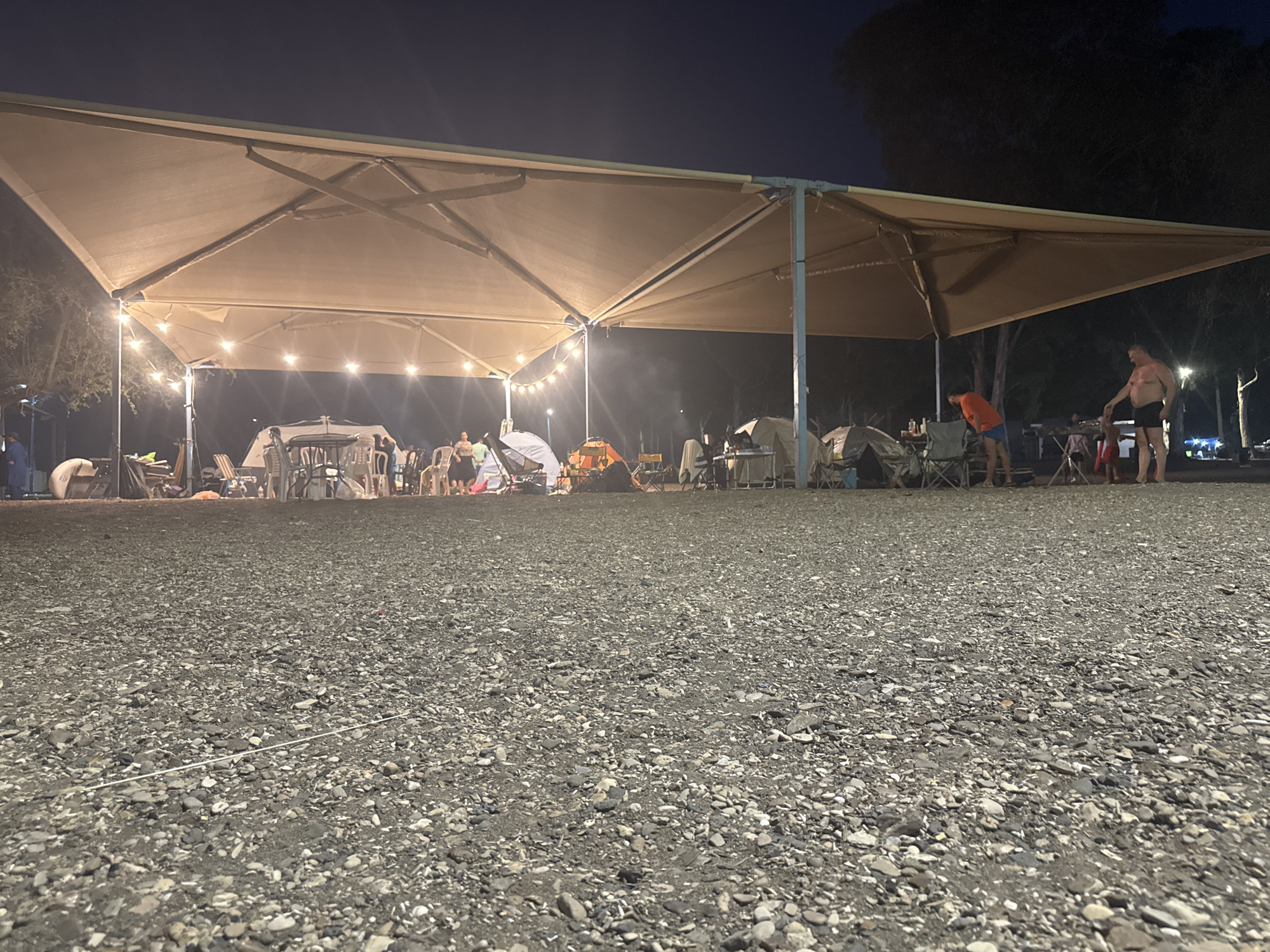 The image depicts a nighttime outdoor gathering under a large, tan awning. The ground is covered with dark gravel. Several tents, camping chairs, and tables are visible beneath the awning. String lights illuminate the area, casting a warm glow.
In the mid-ground, several people are present. On the right side, a shirtless man wearing black shorts stands with his arm extended towards another person. To his left, a person in an orange shirt and blue shorts is bending over. In the background, other individuals are visible, some sitting and some standing, near tents and a table. A white tent is prominent on the left.
The environment appears to be a campsite or an outdoor event space. The dark sky suggests it is late evening or night. The location is identified as the Golan Regional Council, Israel.