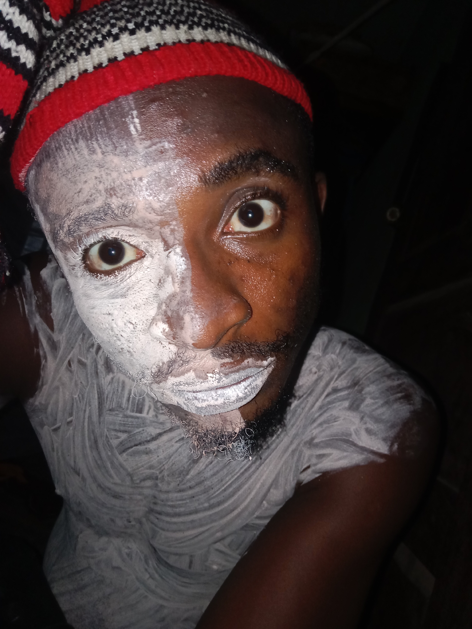 A close-up portrait shows a person with dark skin, wearing a striped red, white, and black knit cap. White paste or paint covers the left side of their face and extends onto their chest and clothing in swirling patterns. The right side of their face is uncovered, revealing their natural skin tone. Their eyes are wide and appear startled or surprised. The background is dark and indistinct. The location is Unknown, Unknown.