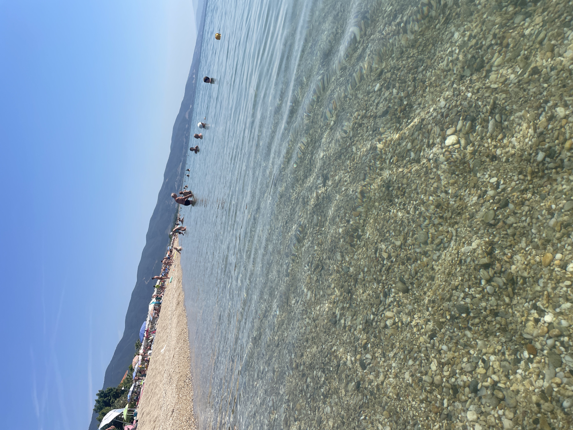 The image captures a sunny, clear day at a pebbly beach in Nea Vrasna, Greece. The foreground features exceptionally clear, shallow seawater, providing an unobstructed view of the seabed composed of multi-sized, multi-colored pebbles and small stones. Gentle ripples on the water surface create subtle distortions of the submerged gravel. In the mid-ground, numerous individuals are present; several people are partially submerged in the water, with their heads and upper torsos visible, engaging in swimming or wading. A yellow buoy is visible floating further out to the right. The pebbly beach itself is populated with multiple individuals, and lined with an array of colorful beach umbrellas (including red, blue, green, and white) and sun loungers. A dense line of green trees is visible at the left edge of the beach. The background consists of a long range of dark, undulating mountains, extending along the coastline and receding into a faint haze towards the horizon. The sky is uniformly clear and bright blue, with no visible clouds.