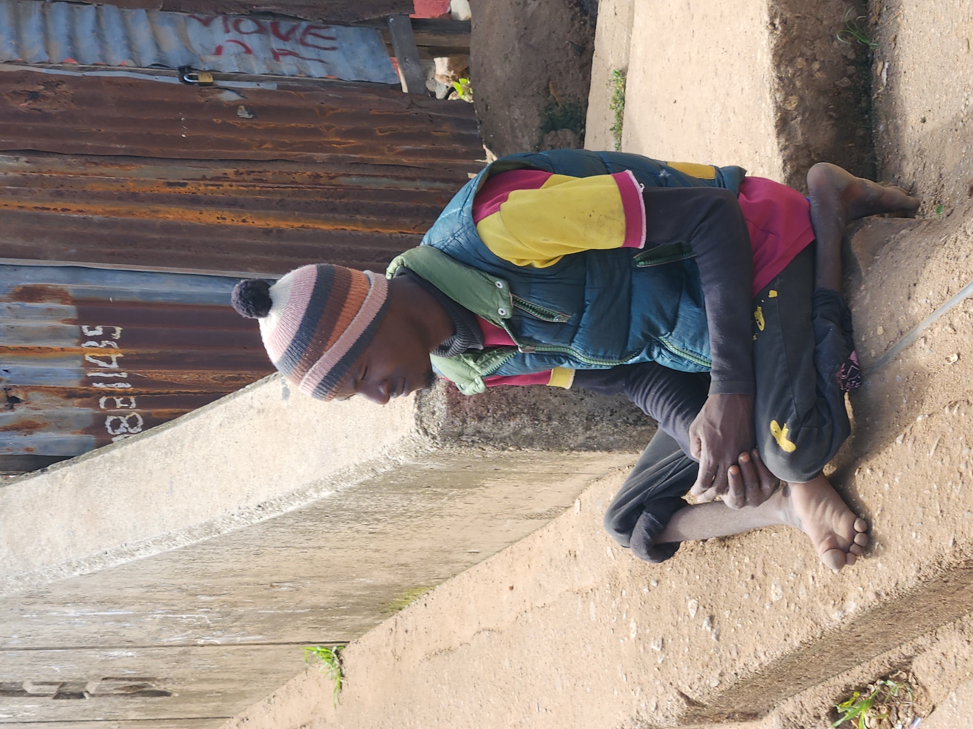 A young Black male wearing a striped beanie, a maroon and yellow long-sleeved shirt, and a teal vest sits on a concrete ledge in Jos, Nigeria. He is barefoot, with his hands resting on his lap. Behind him, a corrugated metal structure with visible rust and graffiti, including the numbers "881145", forms a backdrop. The ground is a mix of dirt and gravel, with small patches of green vegetation. A concrete barrier runs along the left side of the image.