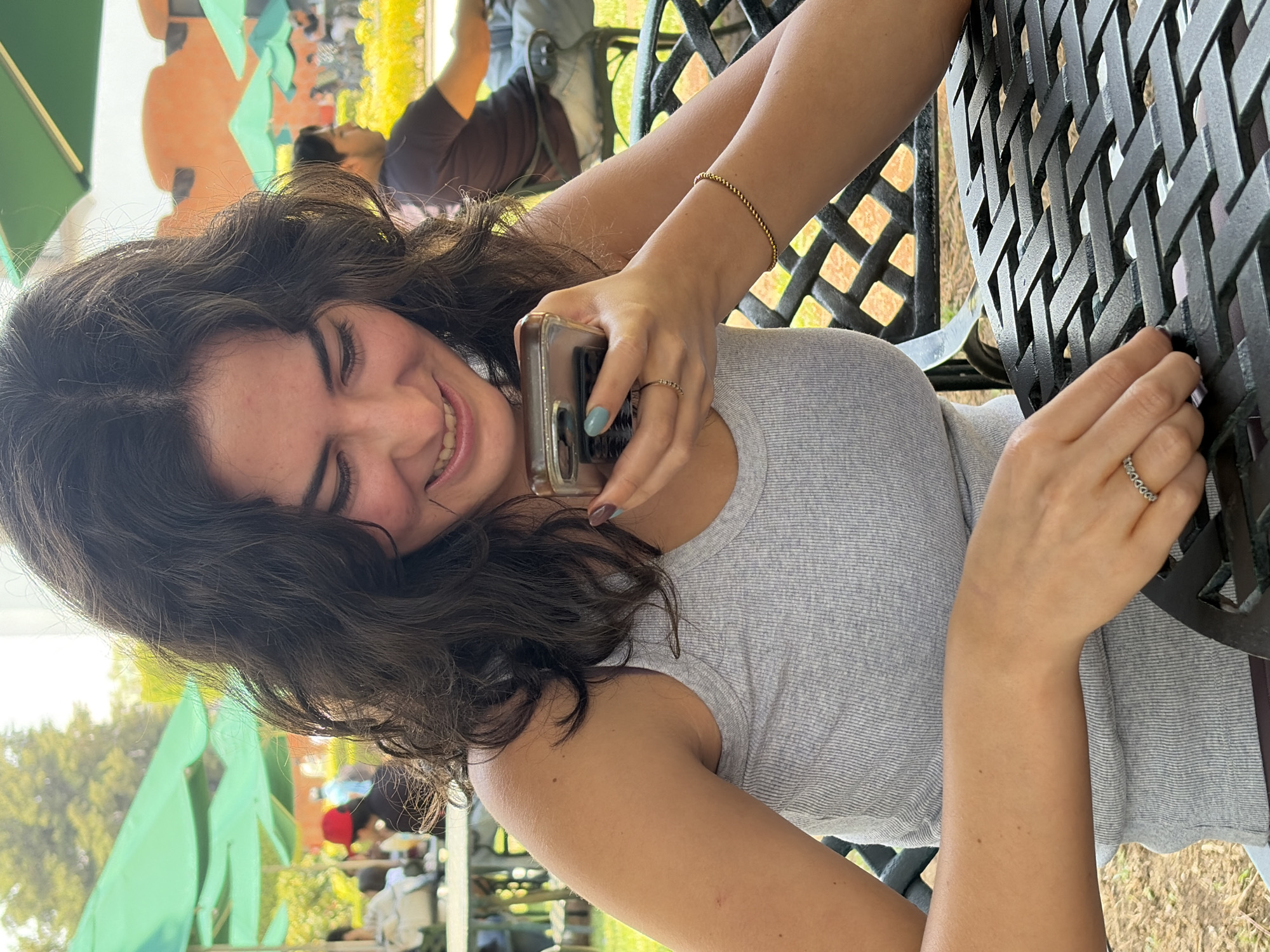 A woman is smiling and holding a cell phone at an outdoor table. She is wearing a grey tank top and a gold beaded bracelet on her left wrist and a ring on the ring finger of her right hand. The table is made of a black metal lattice. In the background, there are other people, green umbrellas, and trees. The location is San Andrés Cholula, Mexico.