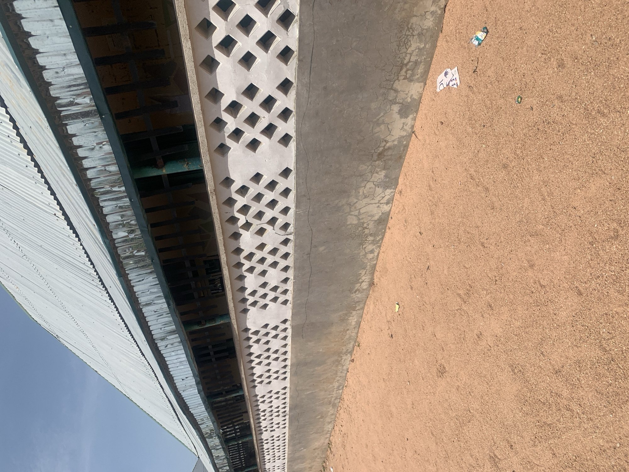 In Jauro Yinu, Nigeria, this image captures the exterior of a building with a corrugated metal roof and a decorative concrete wall. The wall features a repeating pattern of diamond-shaped openings. Below this, a solid concrete base extends to the ground, which is covered in reddish-brown sand. Some litter, including crumpled paper and a small green object, is scattered on the sand near the wall. The sky is clear and blue.