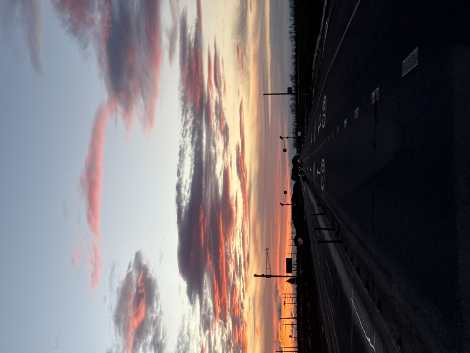 The image displays a wide, multi-lane highway at either sunrise or sunset, characterized by a dramatic and colorful sky. The perspective is looking down the road as it stretches into the distance.
The upper two-thirds of the image are dominated by the sky, filled with long, stretched-out cloud formations. These clouds are painted with vibrant hues of red, orange, and pink, particularly closer to the horizon, which contrasts with darker grey and purple sections within the cloud masses. Higher in the sky, the colors transition to softer yellows and oranges near the horizon, fading into a pale blue and light grey as the sky extends upwards. The lighting suggests a low sun, casting warm tones on the underside of the clouds.
The lower third of the image shows a dark asphalt highway, appearing wide with multiple lanes. Clear white road markings are visible on the surface. Two large, white numbers "60" are prominently painted on the road, indicating a speed limit or lane designation. Additionally, white dashed lines separate the individual lanes.
Along the right side of the highway, a metal guardrail or barrier runs parallel to the road's edge. In the mid-ground, a series of dark silhouettes against the illuminated sky reveal utility poles, streetlights, or gantry structures positioned above or alongside the highway. A faint, dark line on the distant horizon suggests a treeline or landmass. The overall scene evokes a sense of travel or contemplation during a beautiful time of day. No people or vehicles are clearly discernible.